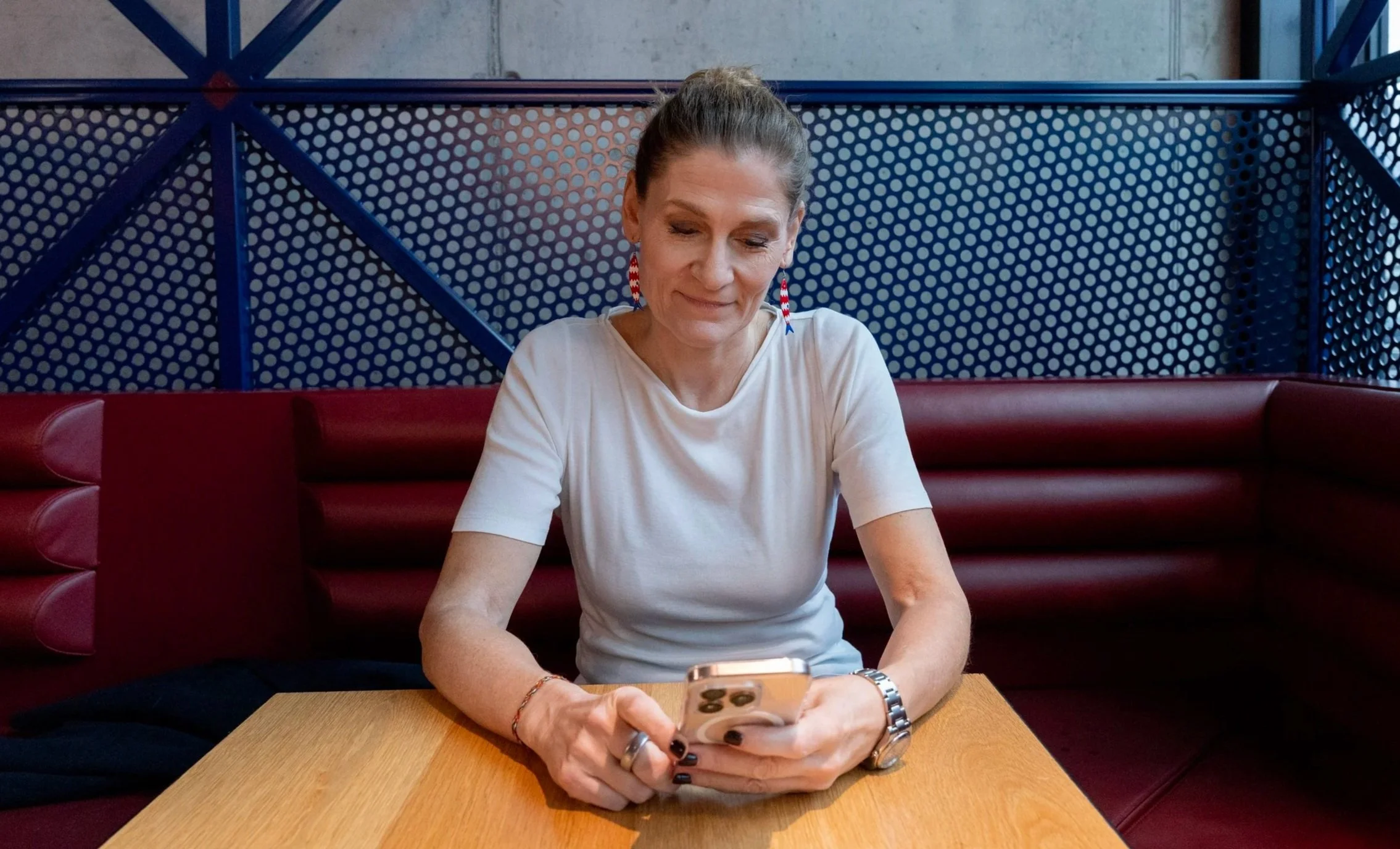 A woman, wearing a white t-shirt and earrings, sitting at a wooden table in a restaurant booth with red cushioned seating, looking at her smartphone.