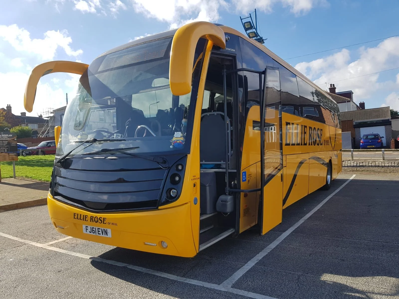 A yellow and black double-decker bus with large banana-shaped handles on the front, parked in a parking lot on a sunny day, with residential houses and a blue sky with clouds in the background.