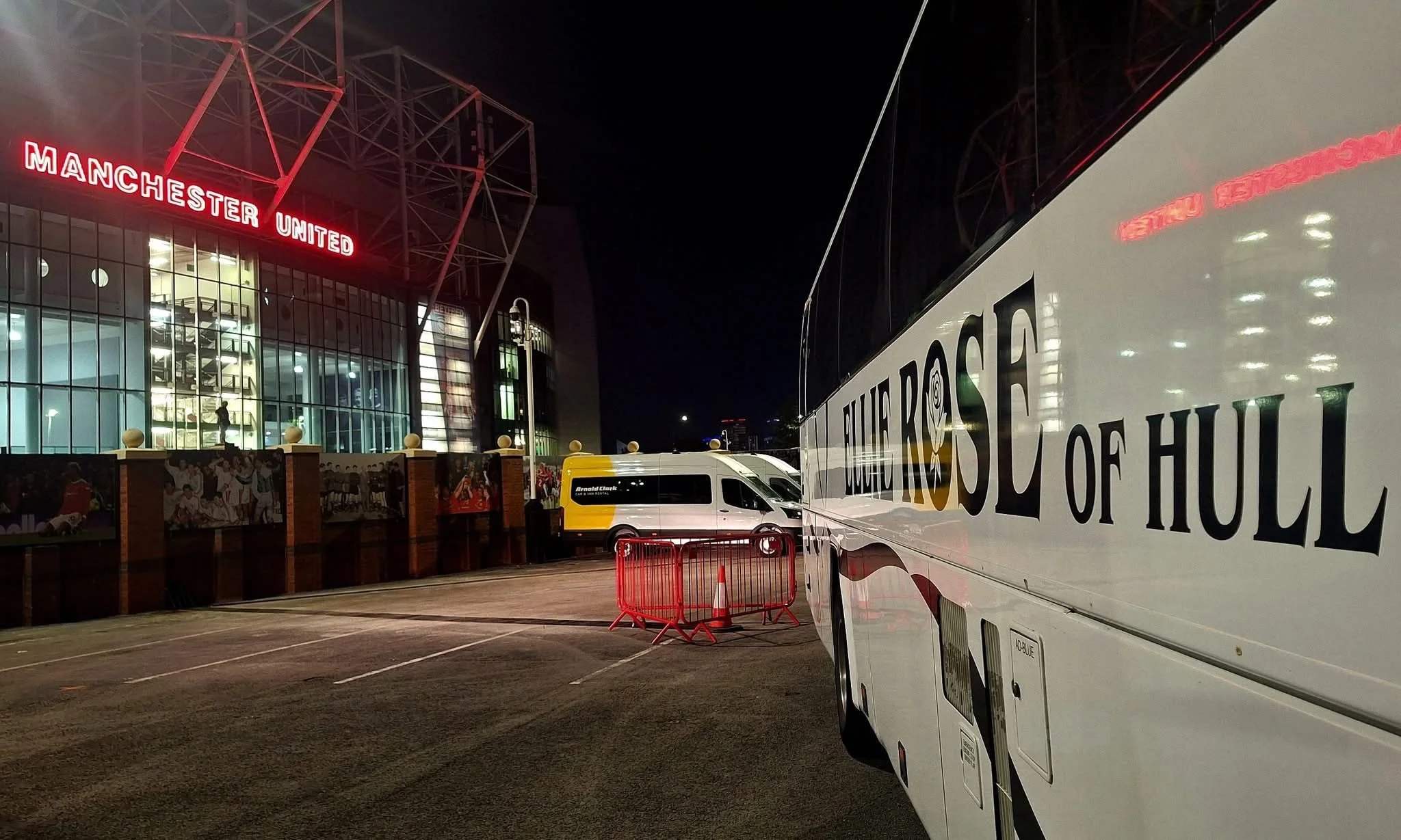 Night scene outside Manchester United stadium with buses parked in the foreground, including one with the words "Blue Rose of HULL" on the side, and the Manchester United sign lit up on the building.