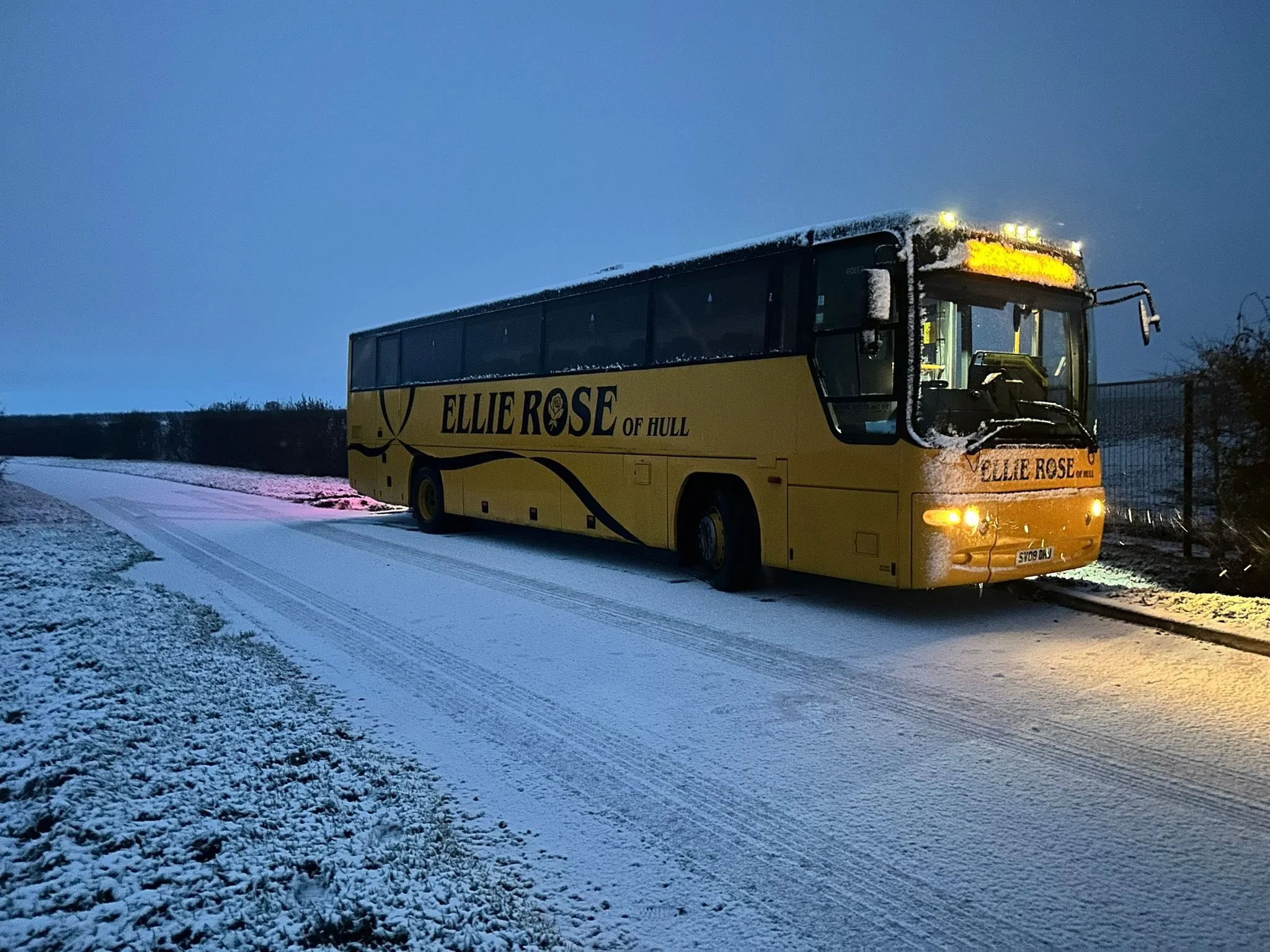 Yellow tour bus with 'Ellie Rose of Hull' written on the side, parked on a snow-covered road during dusk or dawn, with snow on the bus and surrounding area.