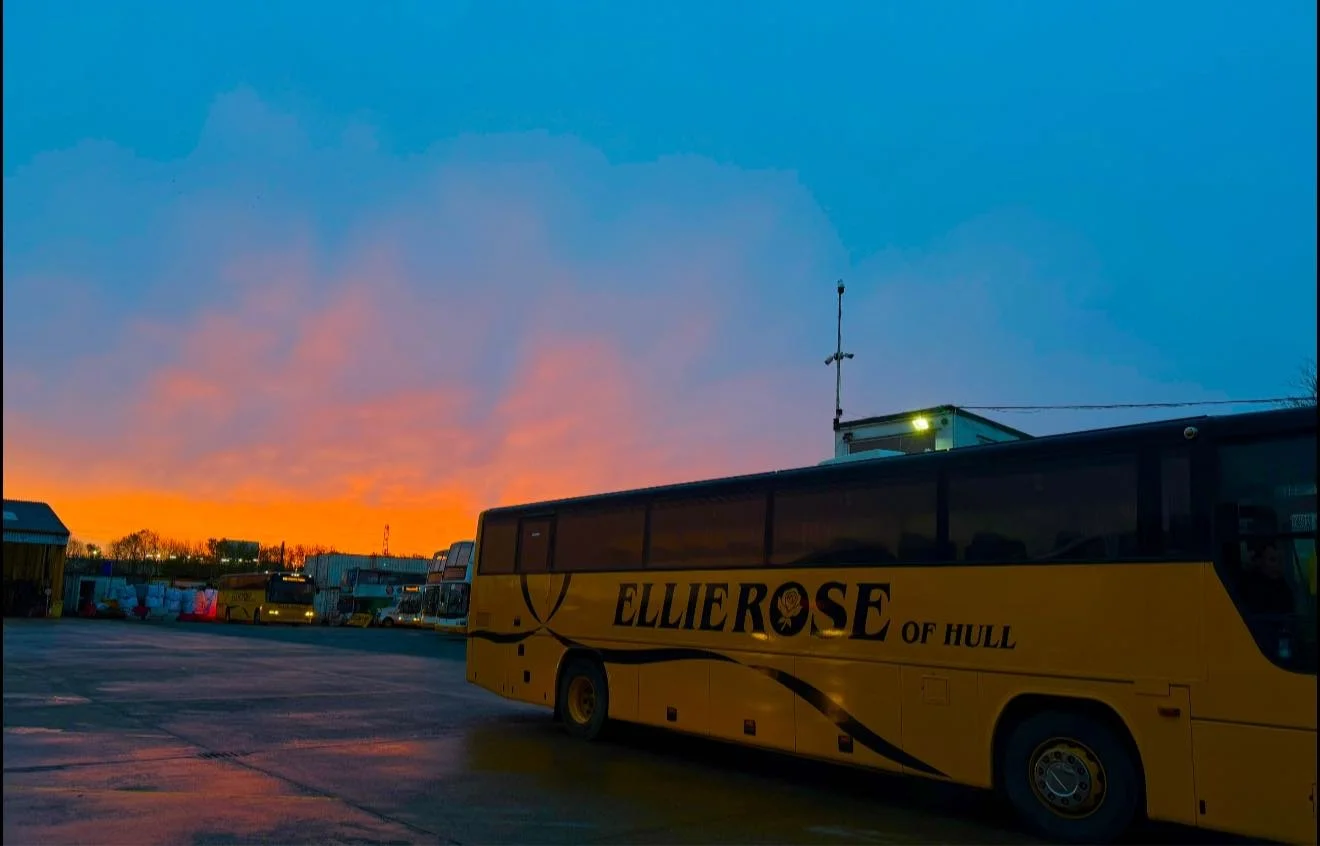 Bus with 'Ellierose of Hull' written on it parked outdoors at sunset, with a colorful sky and other buses in the background.