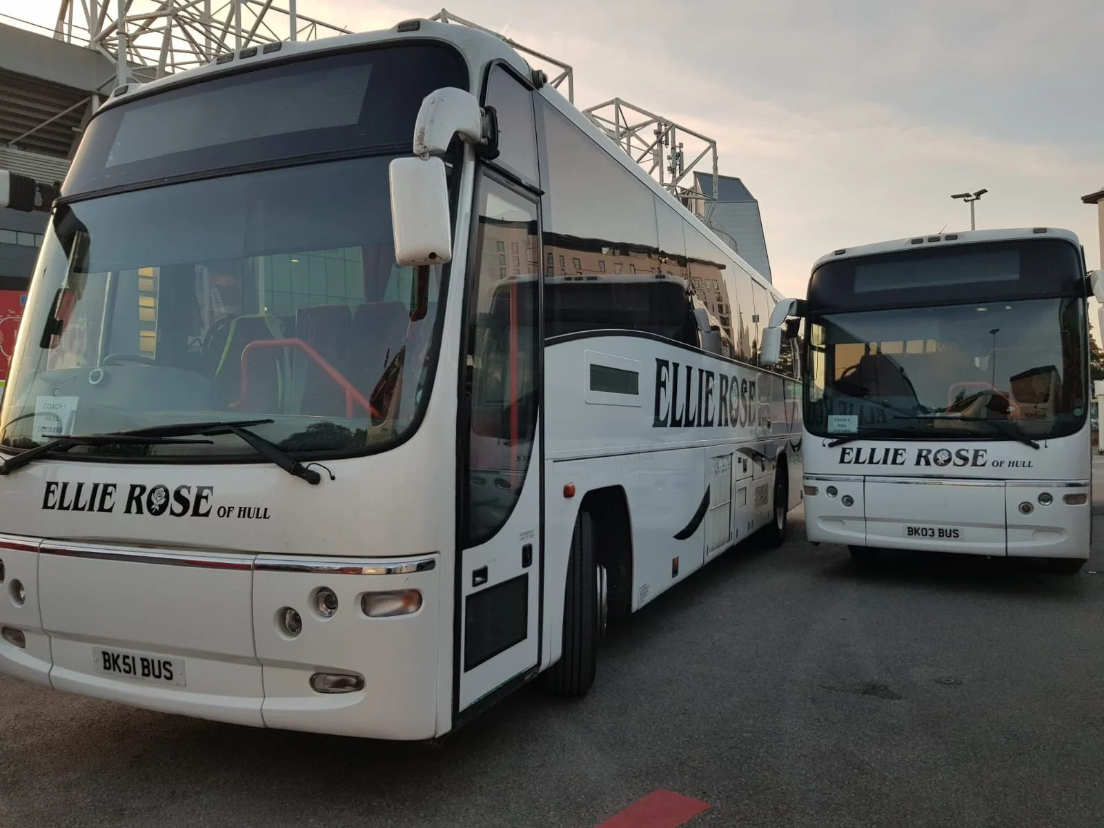 Two white passenger buses with 'Ellie Rose of Hull' on their front and side, parked on a paved lot near a modern building at sunset.