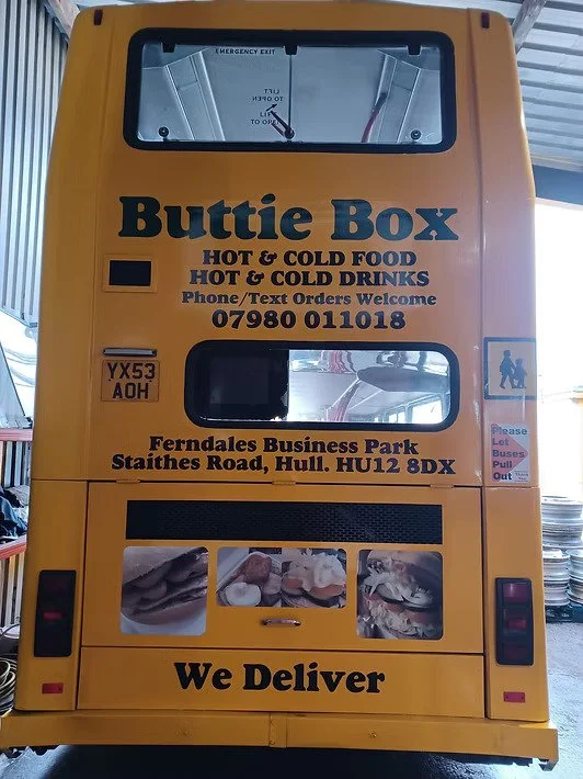 Yellow bus converted into a mobile food delivery unit with a sign that reads 'Buttie Box' offering hot and cold food and drinks. The bus has a large window and a clock at the top. The address is Ferndales Business Park, Staithe Road, Hull, and it includes a contact phone number. There are pictures of sandwiches at the bottom, and the bus is labeled 'We Deliver'.