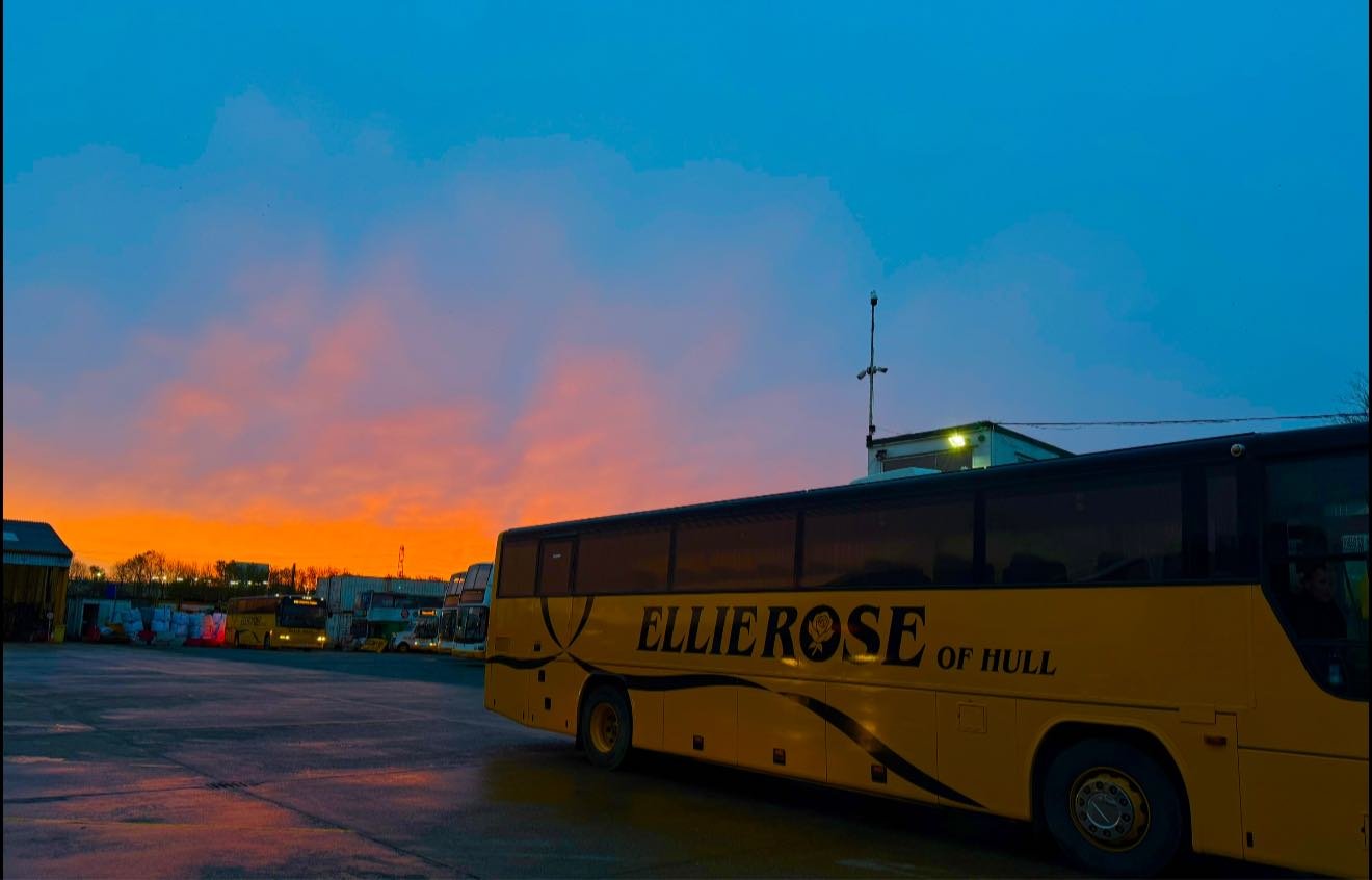 Yellow bus with 'Ellierose of Hull' parked in a lot during sunset, with colorful sky and other buses in the background.