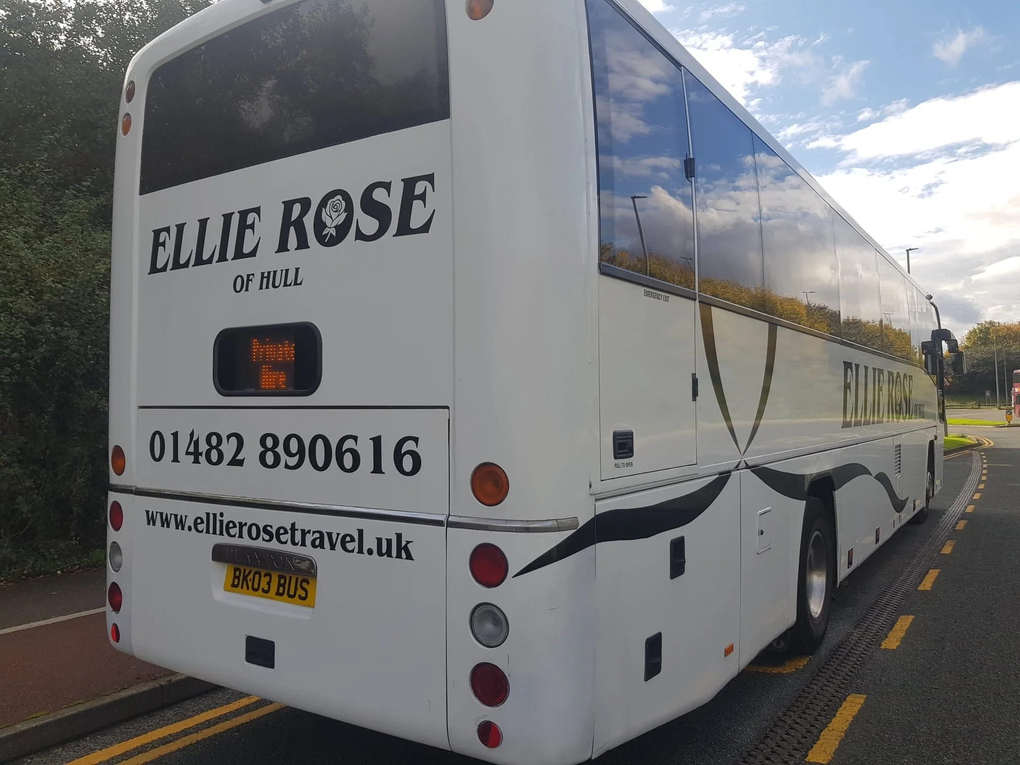 A white Elllie Rose of Hull tour bus parked on a road with trees in the background.