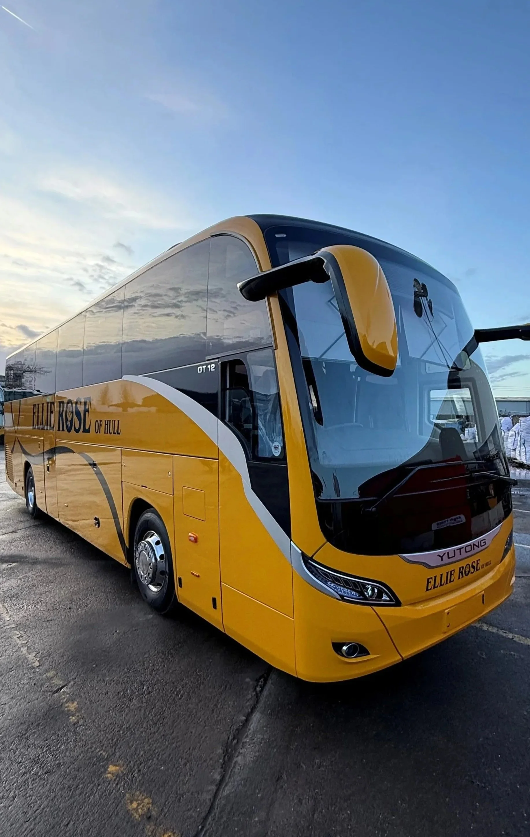 A yellow and black coach bus parked on a paved surface against a sky with clouds at sunset.