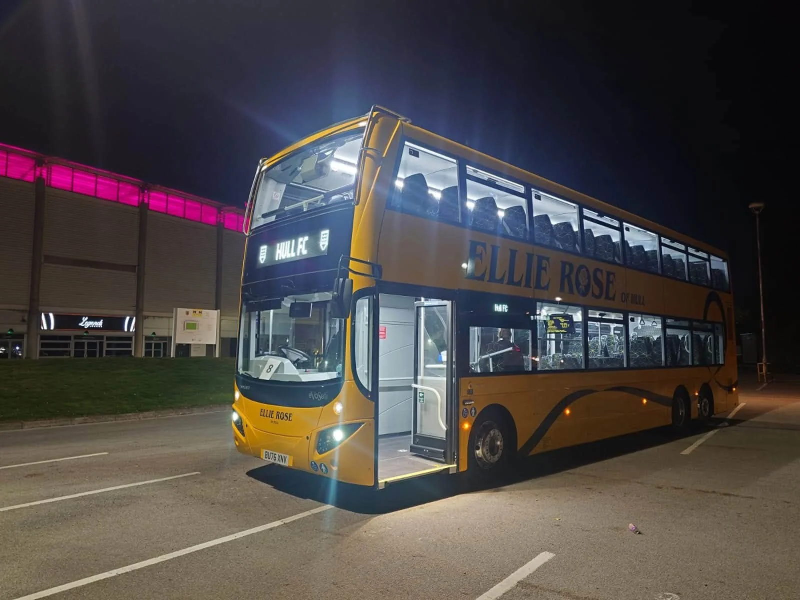 A double-decker bus parked at night with its doors open and interior lights on, displaying route information for HULL FC, with a dark sky and a building with pink lighting in the background.