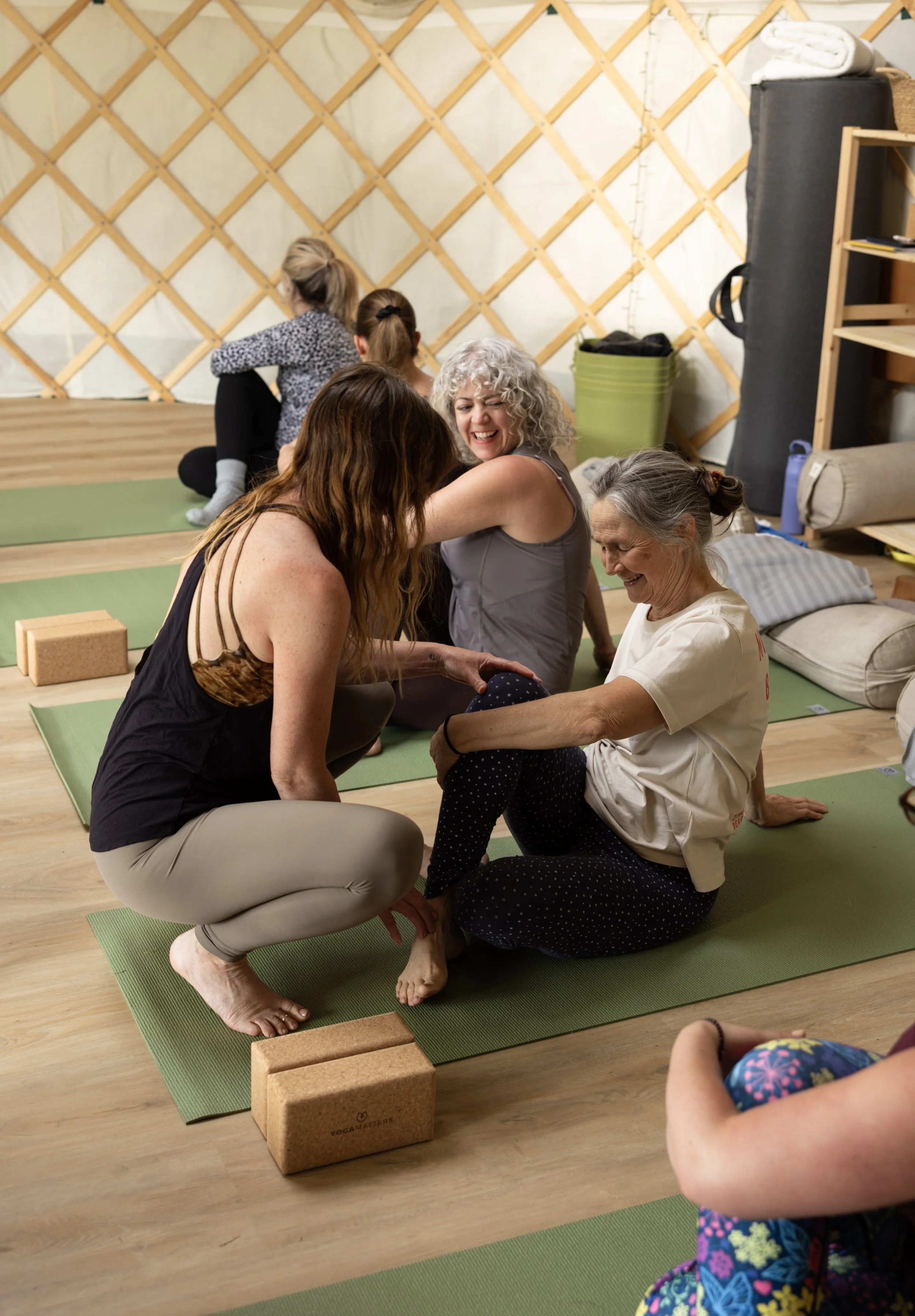 A group of women and children participating in a yoga class in a room with wooden flooring and a lattice wall. The women are sitting on yoga mats practicing a partner pose, with some children in the background. The atmosphere appears joyful and engag