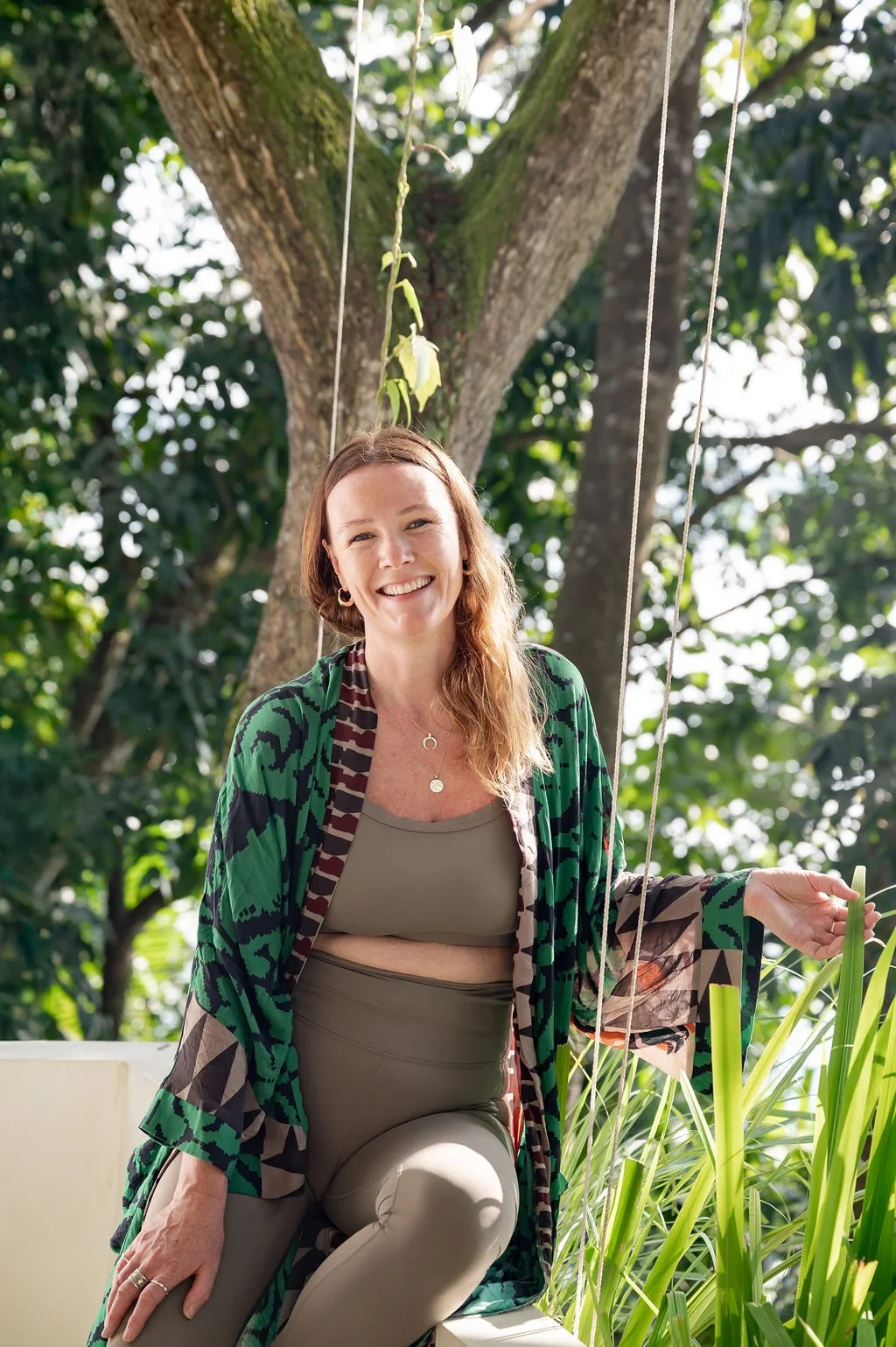 A woman with long, wavy hair smiling outdoors, wearing a green and black patterned jacket over a beige sports bra and beige leggings, sitting on a white surface with greenery and a tree in the background.