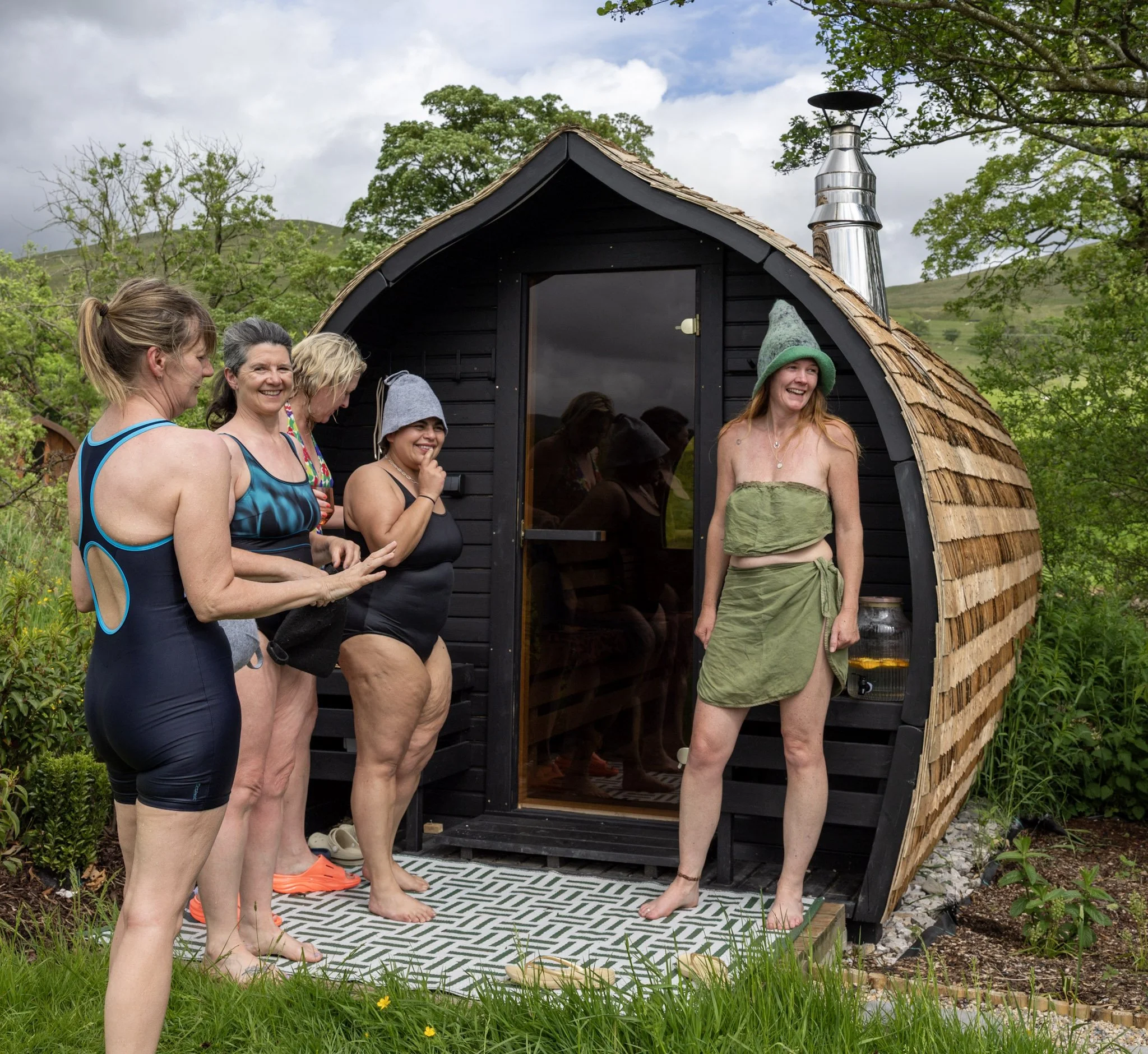 Group of women in swimwear standing outside a small black cabin with a curved roof, surrounded by scenic greenery and trees, under a partly cloudy sky.