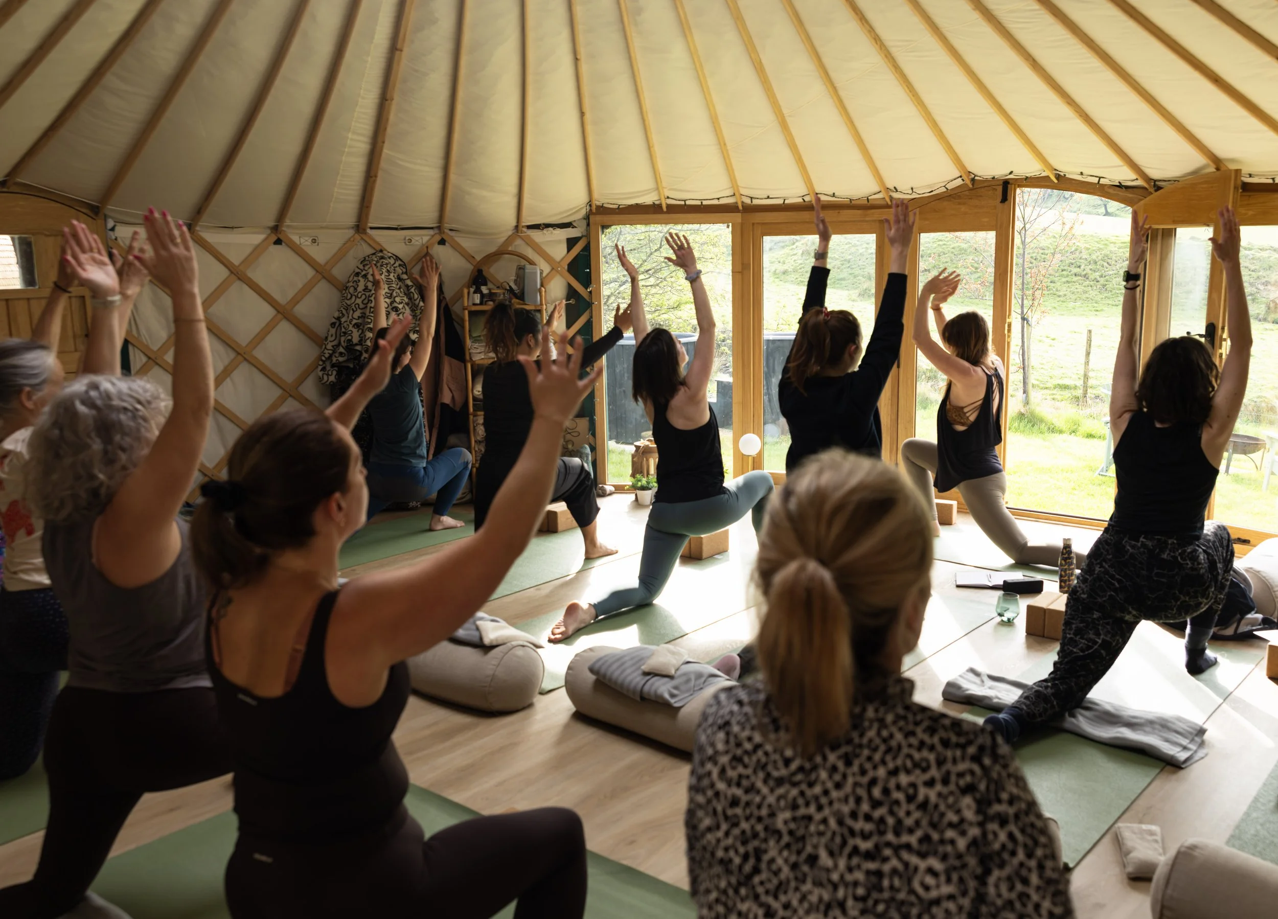 A group of people participating in a yoga class inside a wooden yurt with large windows showing a green outdoor landscape, some individuals are in a warrior pose with arms raised.