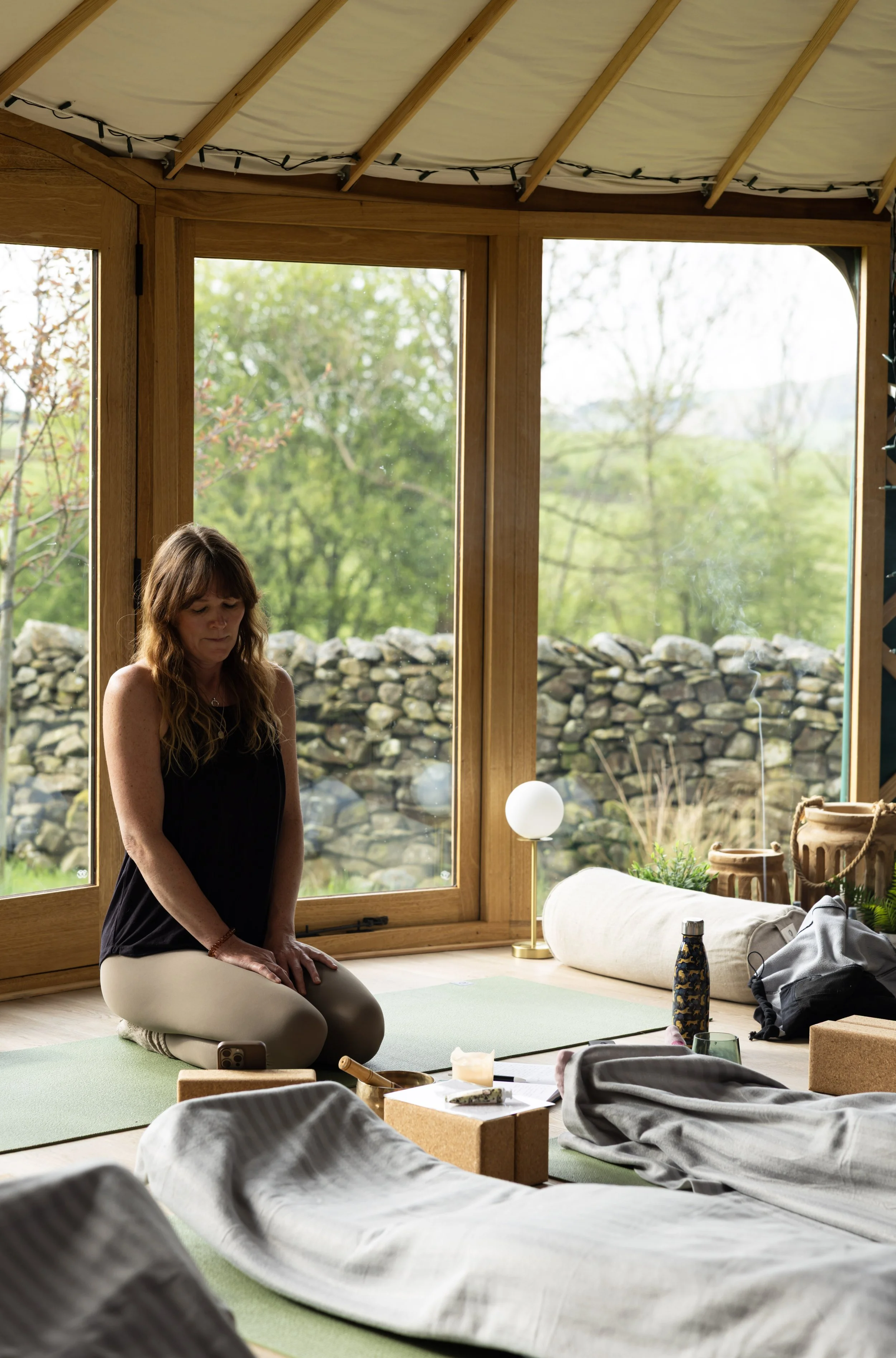 A woman practicing yoga or meditation on a green yoga mat inside a room with large windows overlooking a scenic outdoor landscape with trees and a stone wall. Various items, including a water bottle, notebook, and stationary, are scattered nearby.