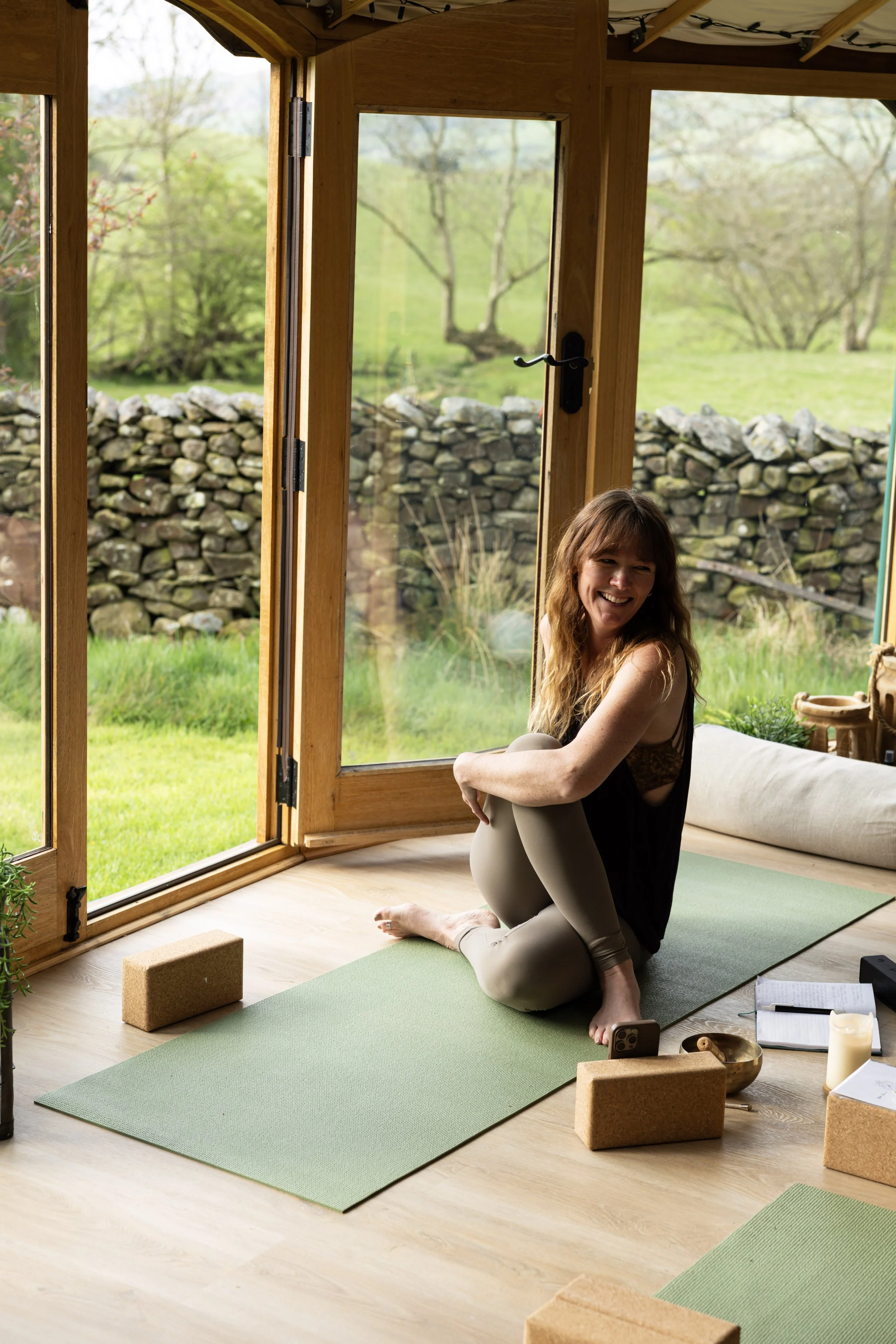 A woman sitting on a yoga mat in a bright room with large glass doors opening to a lush green outdoor landscape. She is smiling and appears relaxed. Yoga props are around her, including yoga blocks, a mat, and small decorative items.
