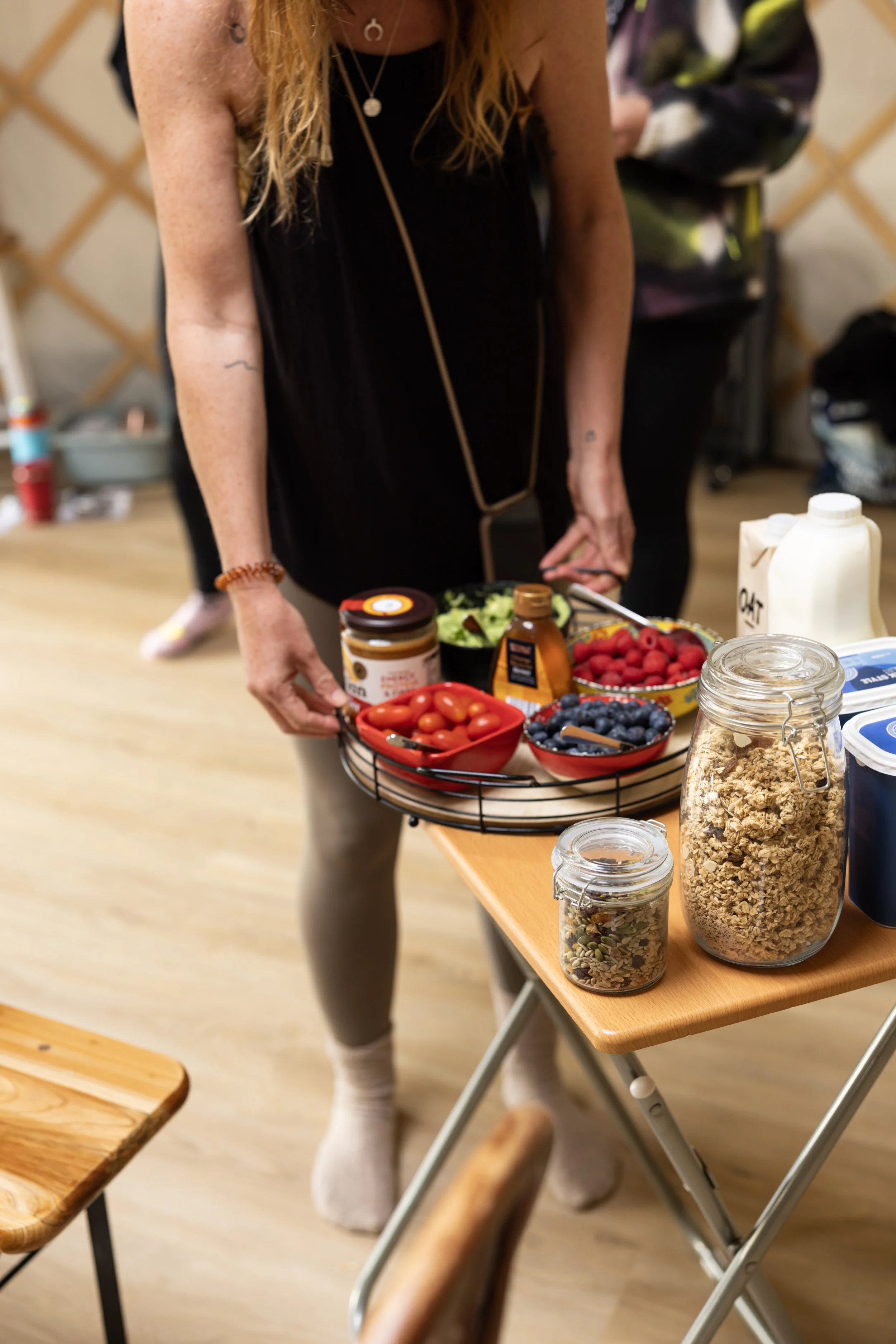 A woman with long blonde hair wearing a black dress and white socks stands at a table with various food items, including berries, granola, and a carton of oats, in a room with a wooden floor and a lattice wall in the background.