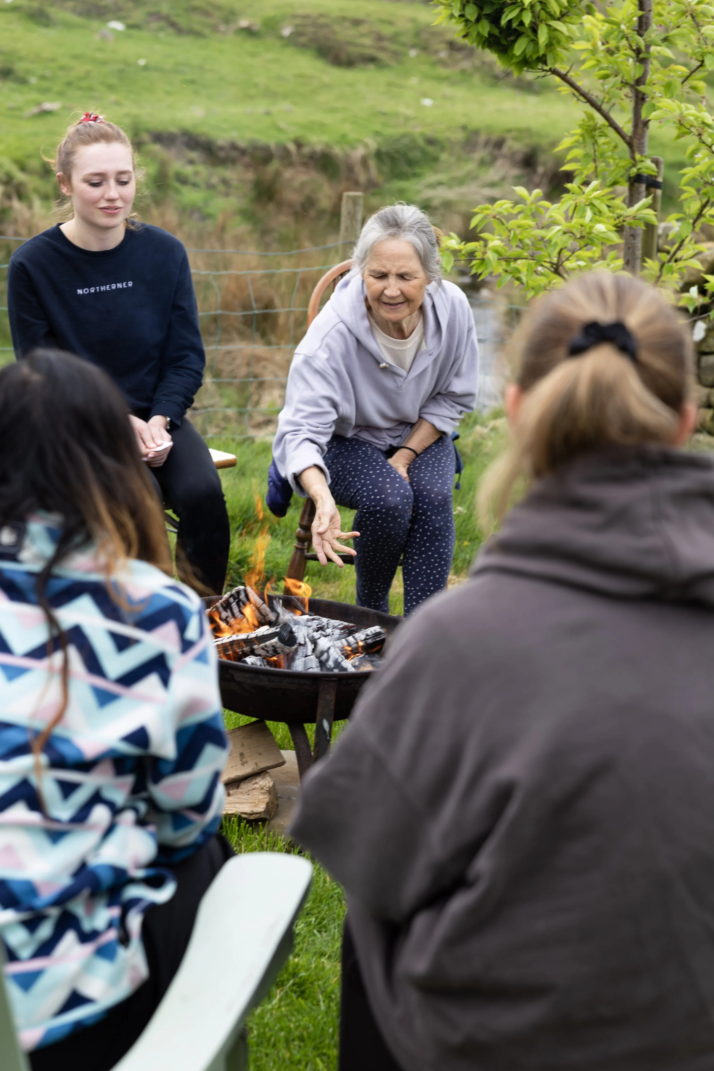 A group of people sitting outdoors around a fire pit, with an elderly woman speaking and gesturing, while others listen in a garden setting.