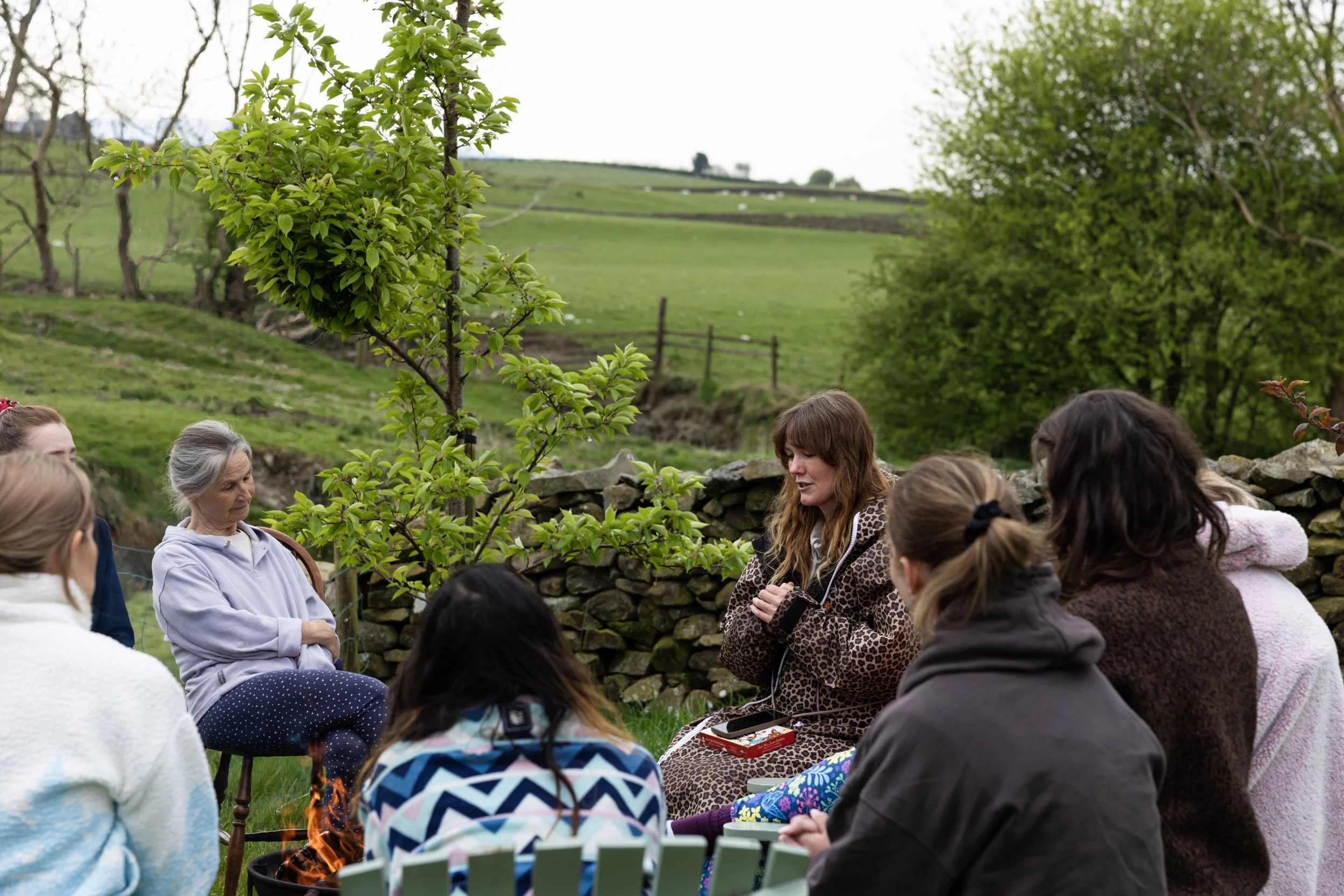 A group of women and girls sitting outdoors in a grassy field with trees and rolling hills in the background, engaged in a discussion or lesson. The scene suggests a casual or community gathering in a rural setting.