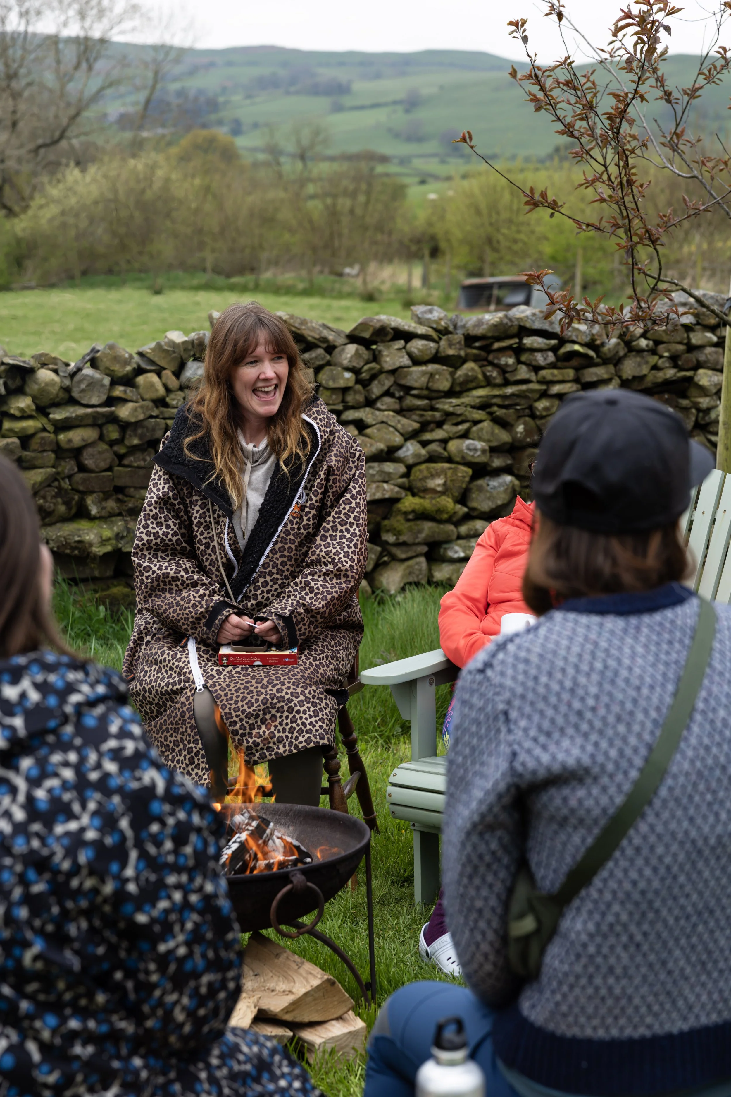 Group of people sitting outdoors around a fire, with a woman laughing, a stone wall, and a scenic background of rolling green hills and trees.