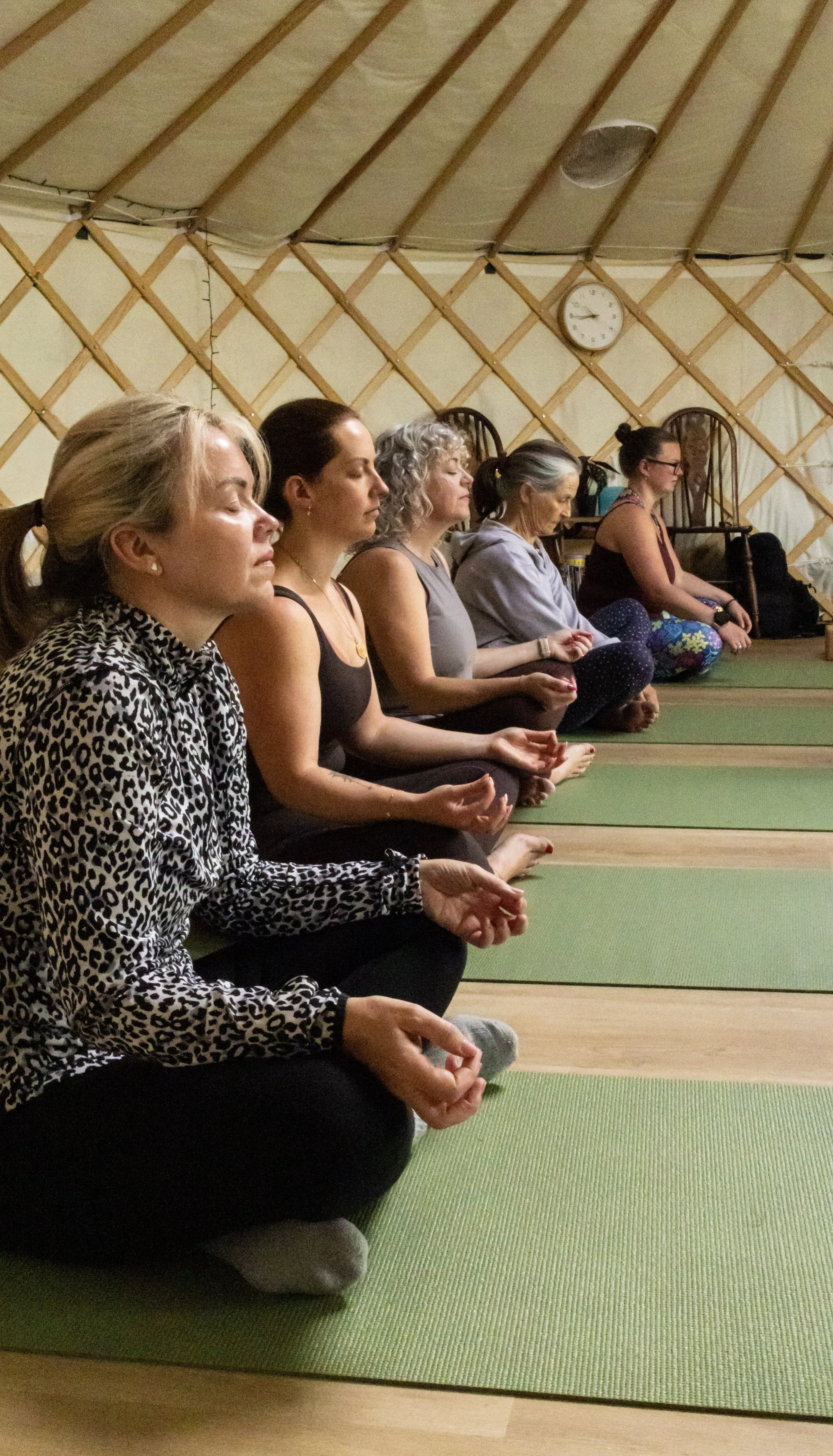 Six women sitting on green yoga mats in a yoga or meditation studio, meditating with eyes closed.