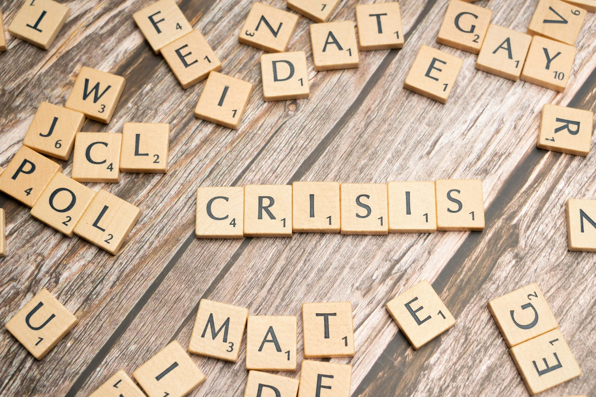 Scrabble tiles spelling out 'CRISIS' on a wooden surface, surrounded by scattered letter tiles.