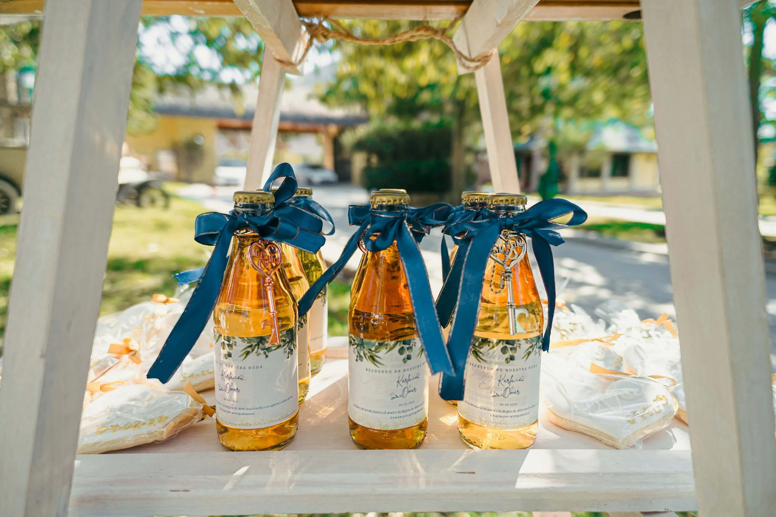 Glass bottles with amber liquid decorated with blue ribbons and placed on a wooden display stand, set outdoors with trees and houses in the background.