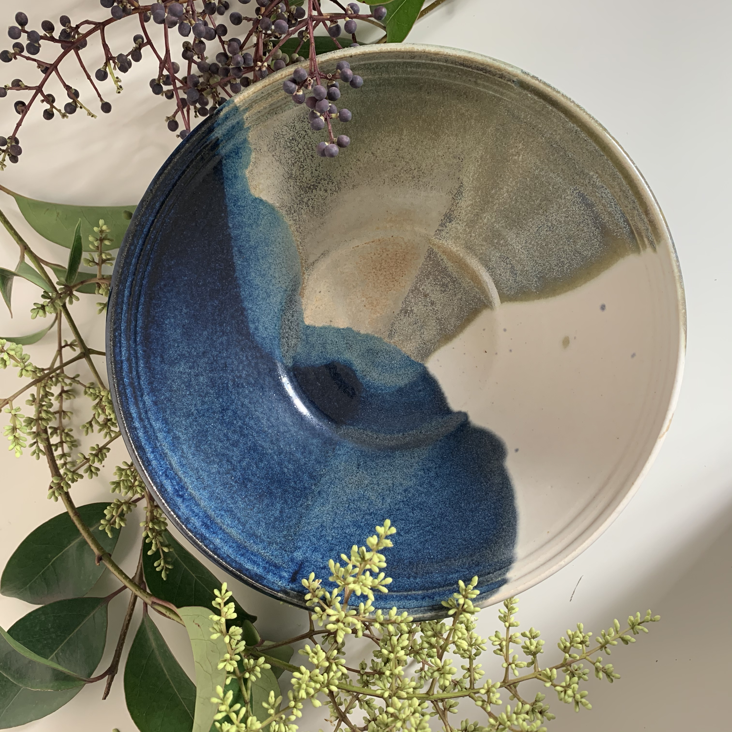 A ceramic bowl with blue, green, and beige glaze inside, surrounded by sprigs of green leaves and small purple berries.