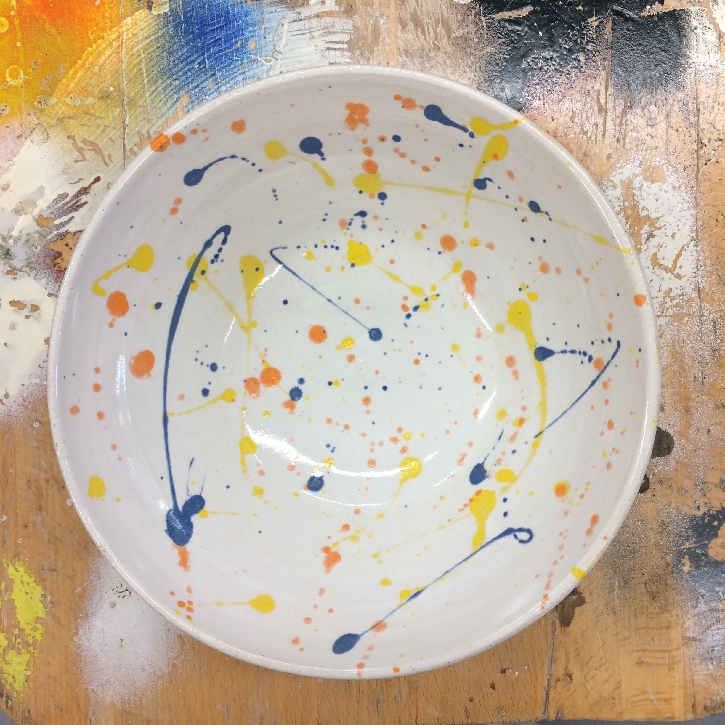 White ceramic plate with a splatter paint design in yellow, blue, and orange, placed on a paint-splattered work surface.