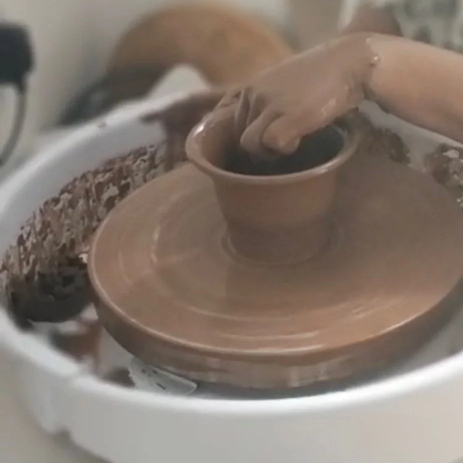 A person shaping a small clay cup on a rotating pottery wheel, using their hand to mold the wet clay.