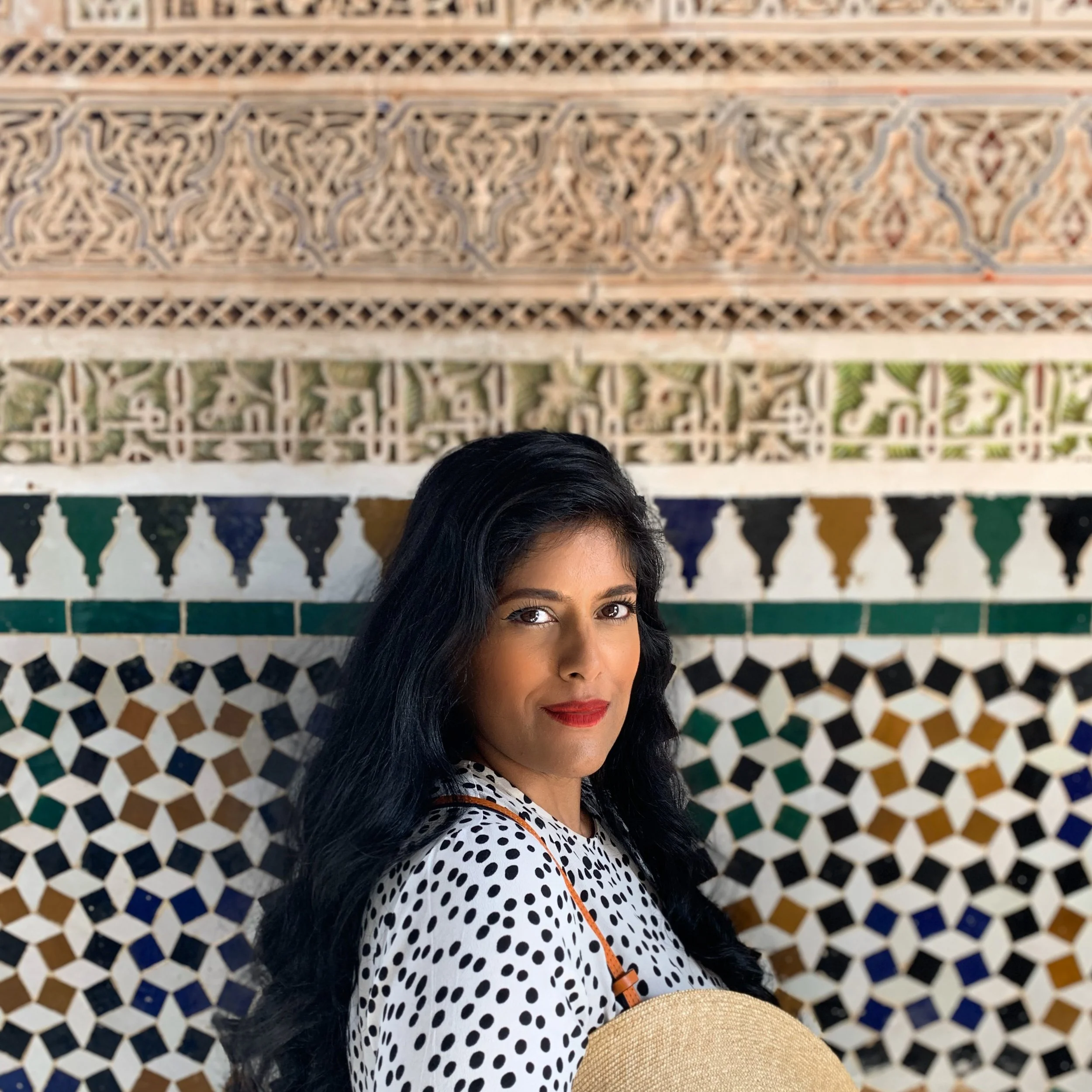 A woman with long black hair, wearing a white shirt with black polka dots, standing in front of a colorful, ornate tiled wall.