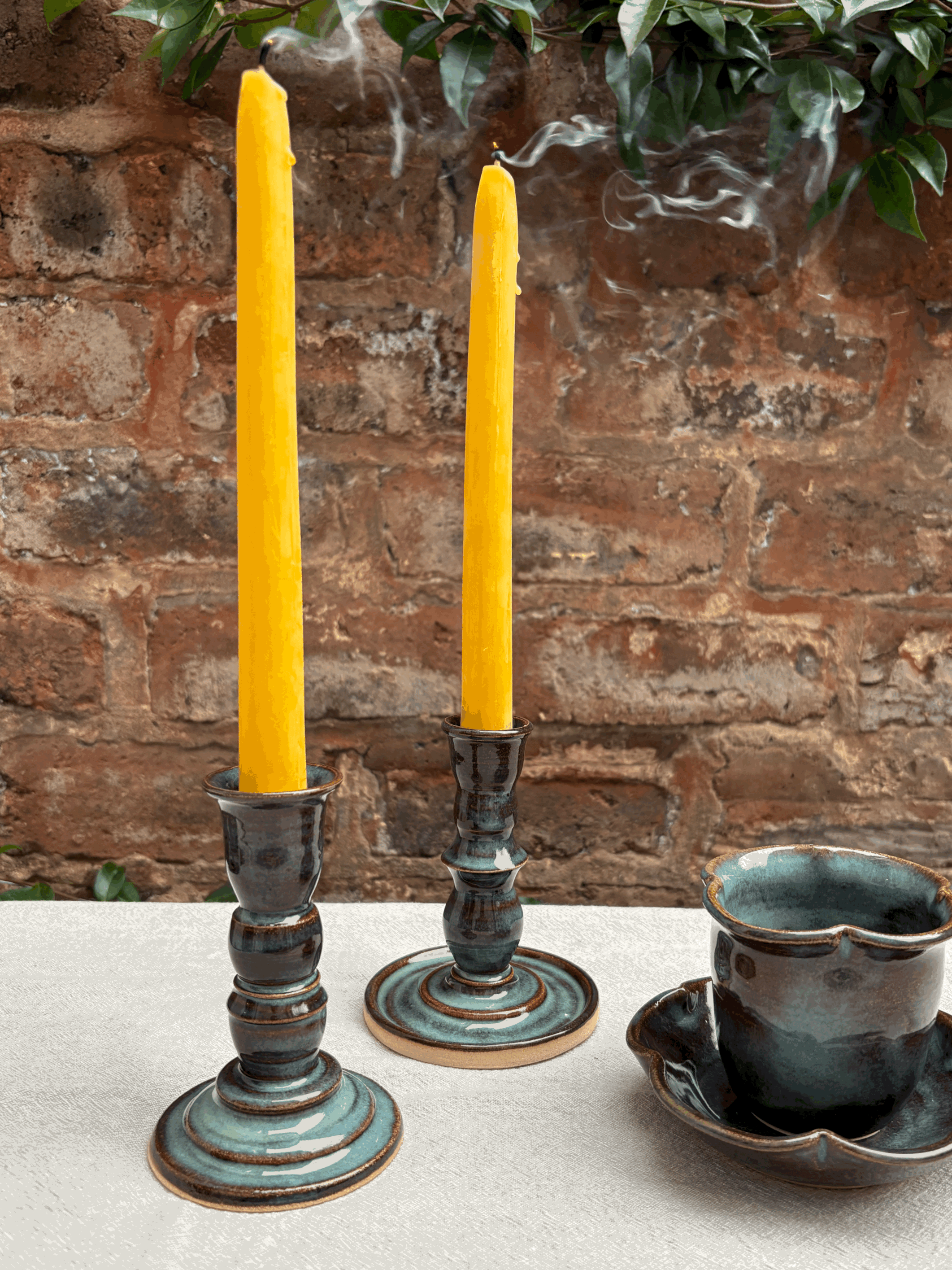 Two yellow candles in ceramic holders and a ceramic cup with saucer on a white table against a brick wall background with greenery.