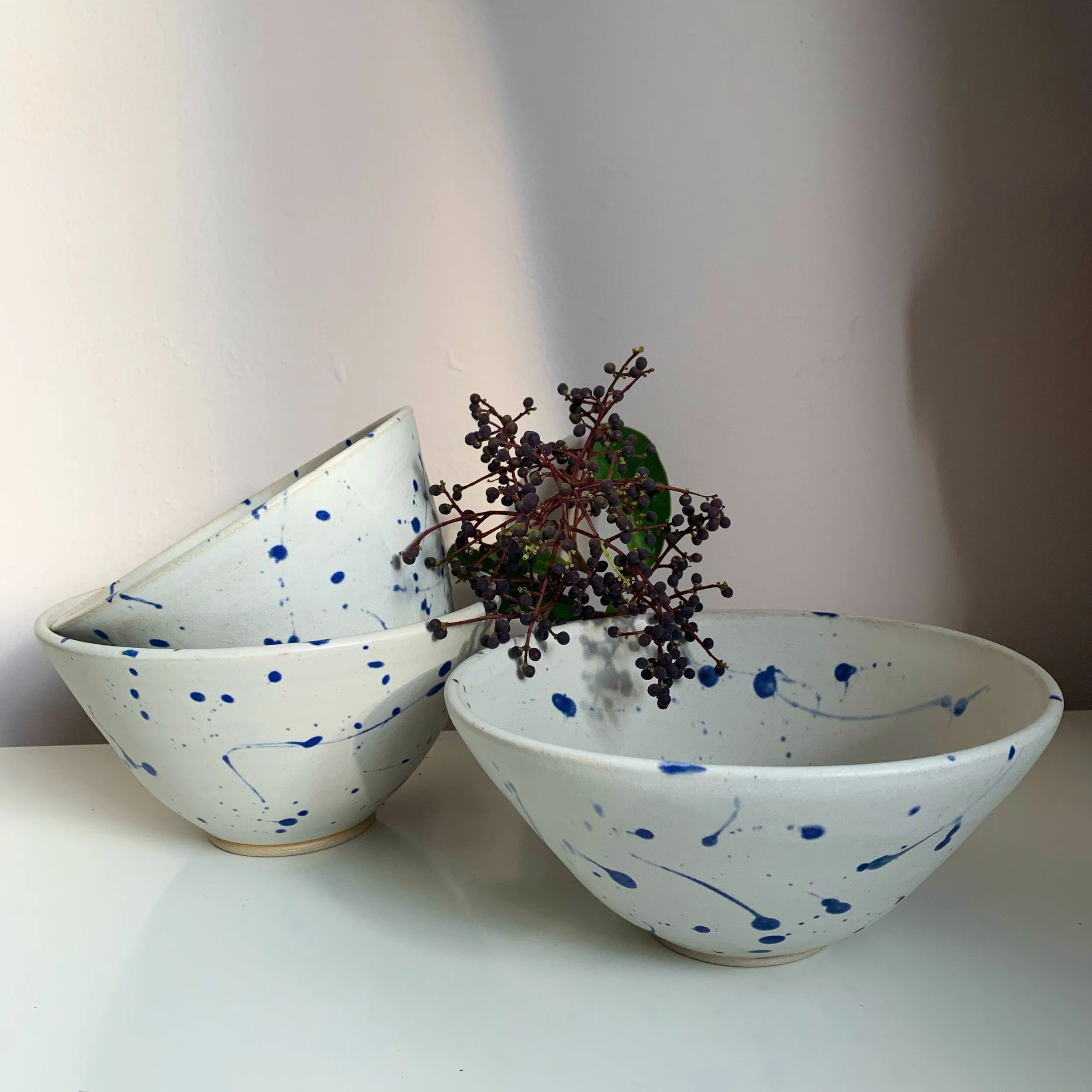 Three white ceramic bowls with blue splash patterns, one stacked inside another, with a small branch of dark purple berries and green leaves on top, on a white surface against a plain wall.