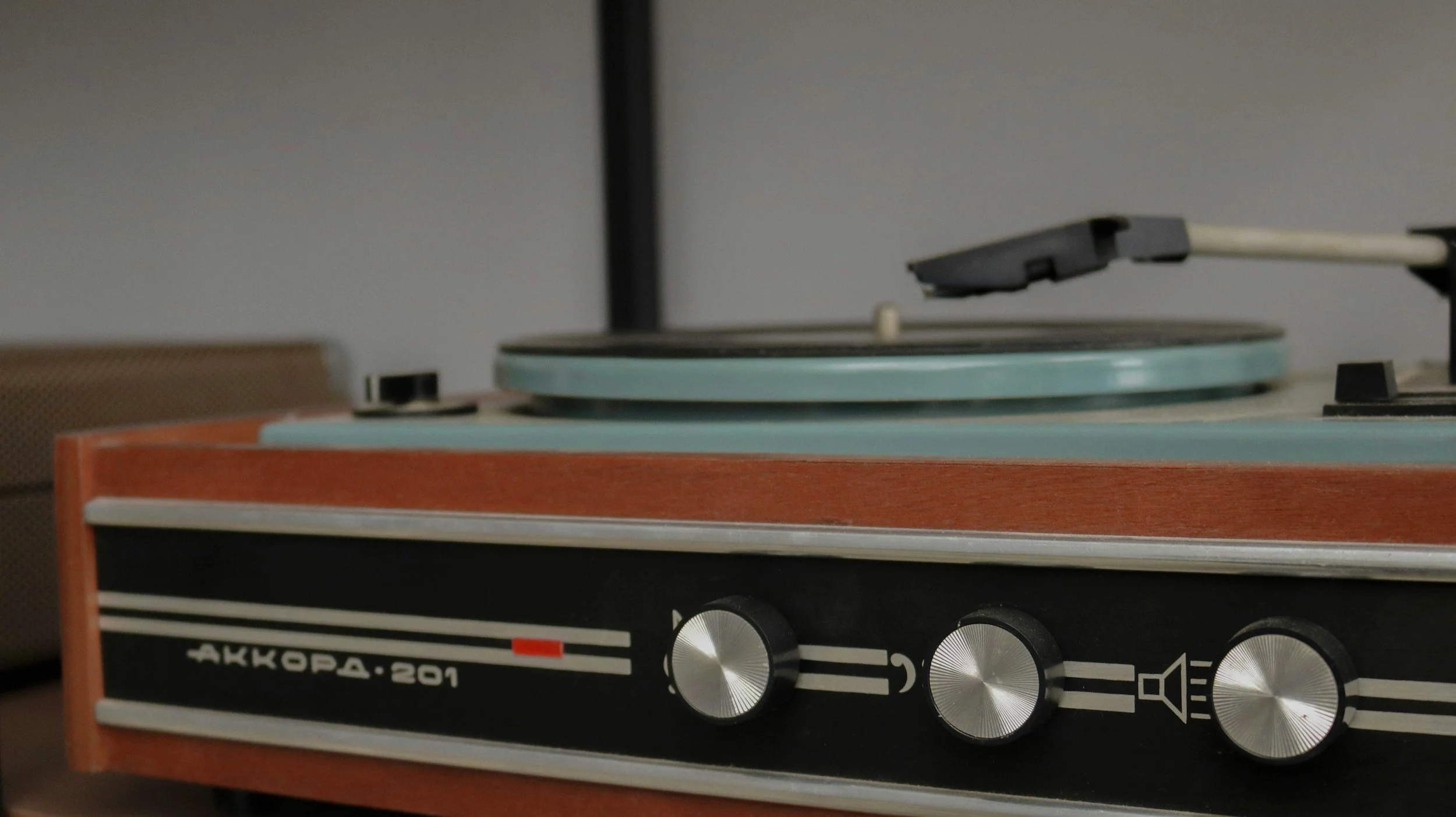 Close-up of a vintage turntable with three silver control knobs on a black panel, brand name 'АККОРД-201' in Cyrillic, and a wooden frame, with a stylus arm and spinning record on the platter.