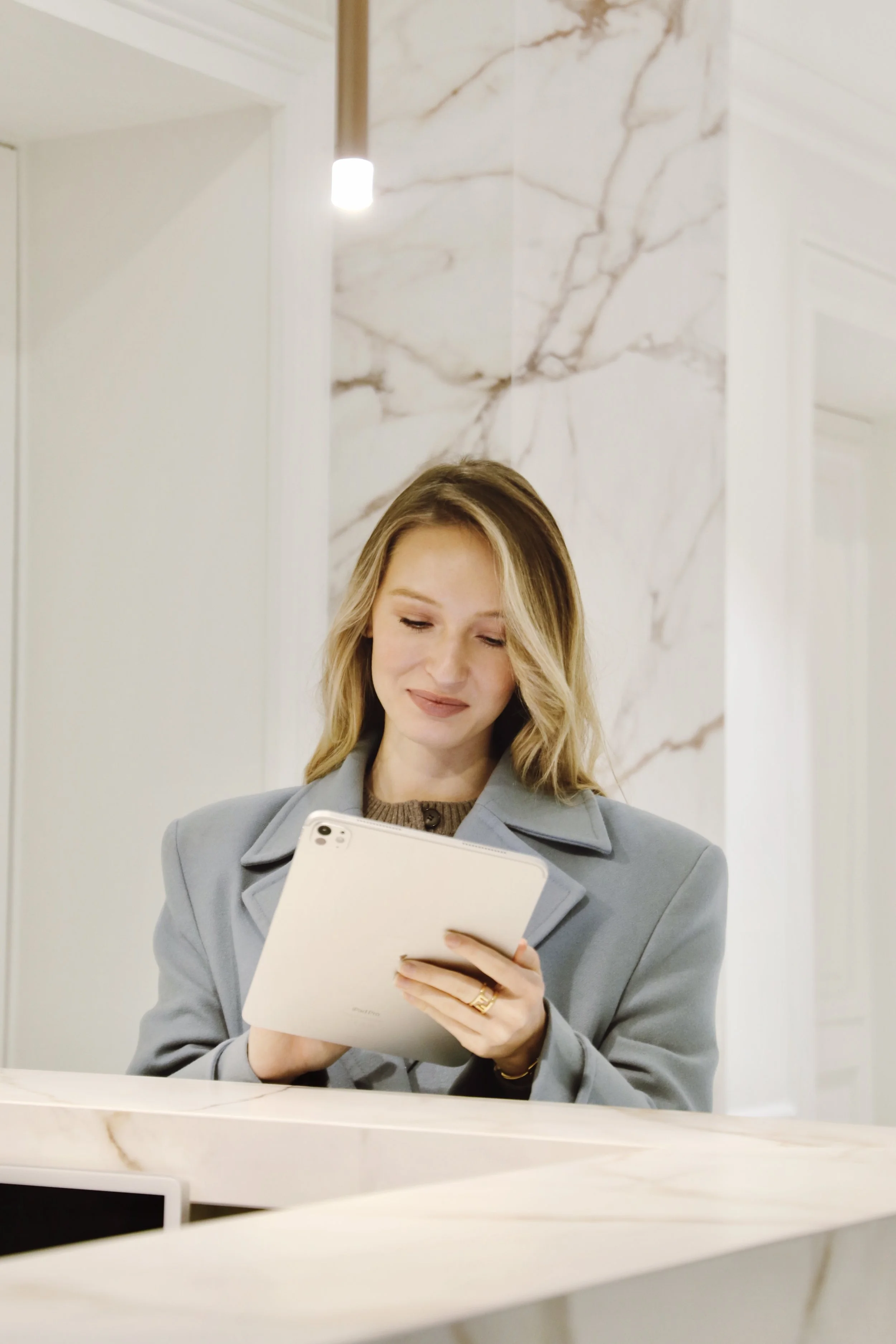 Une femme souriante regarde une tablette ou un smartphone blanc, dans un intérieur élégant avec un mur en marbre blanc et des lumières modernes.