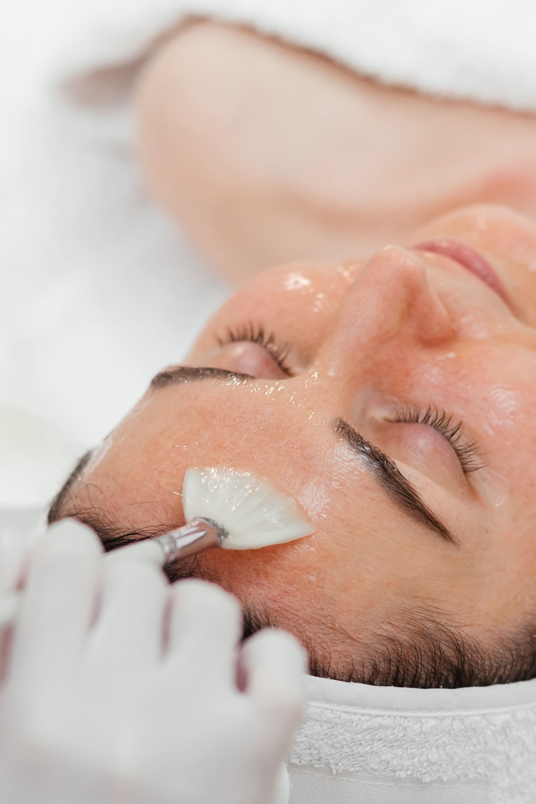 Une personne reçoit un soin du visage, avec un professionnel appliquant une crème ou un masque sur le visage à l'aide d'un pinceau, lors d'une séance de spa ou de soins esthétiques.