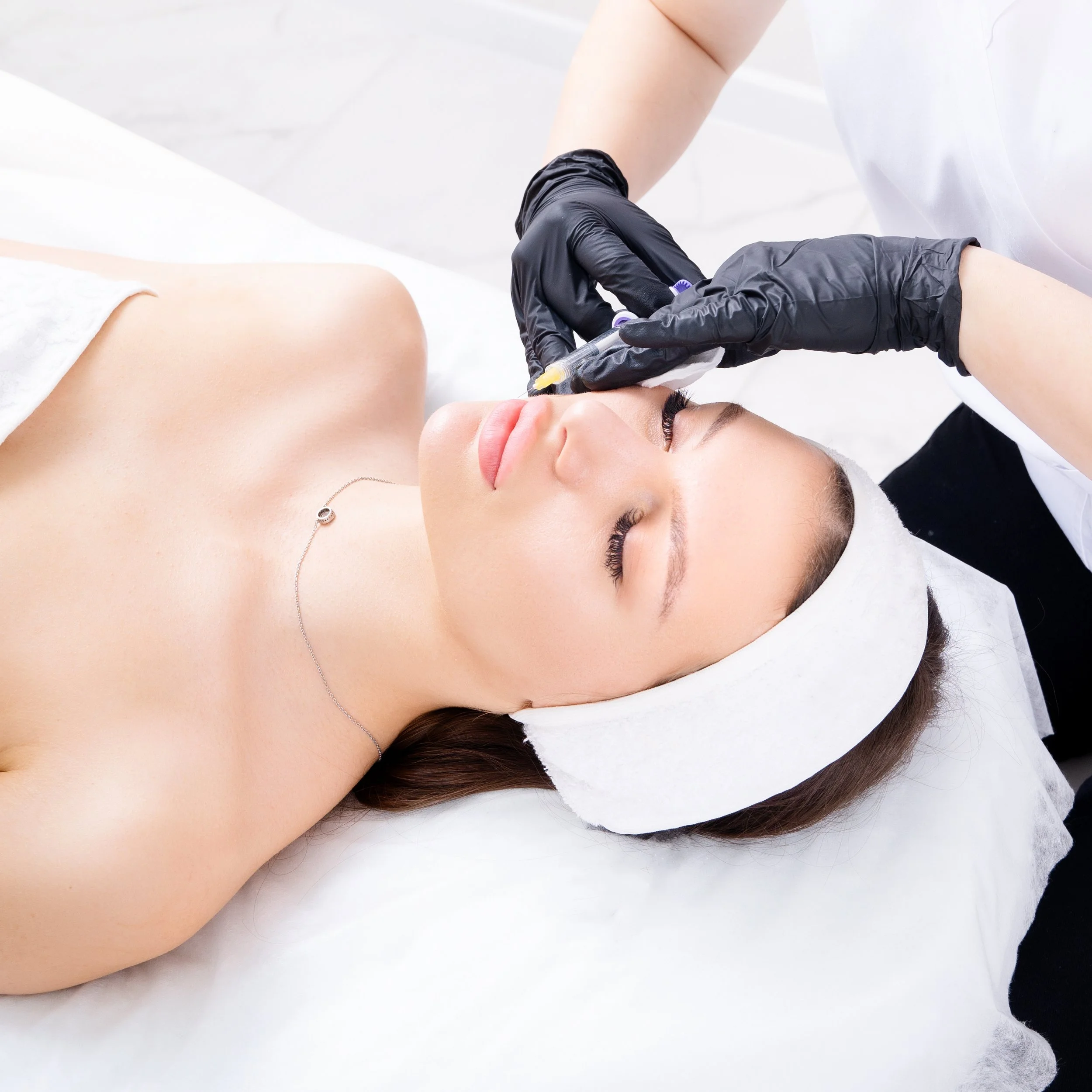 Une femme reçoit une injection dans le visage lors d'un traitement esthétique, allongée avec un bandeau blanc sur la tête.