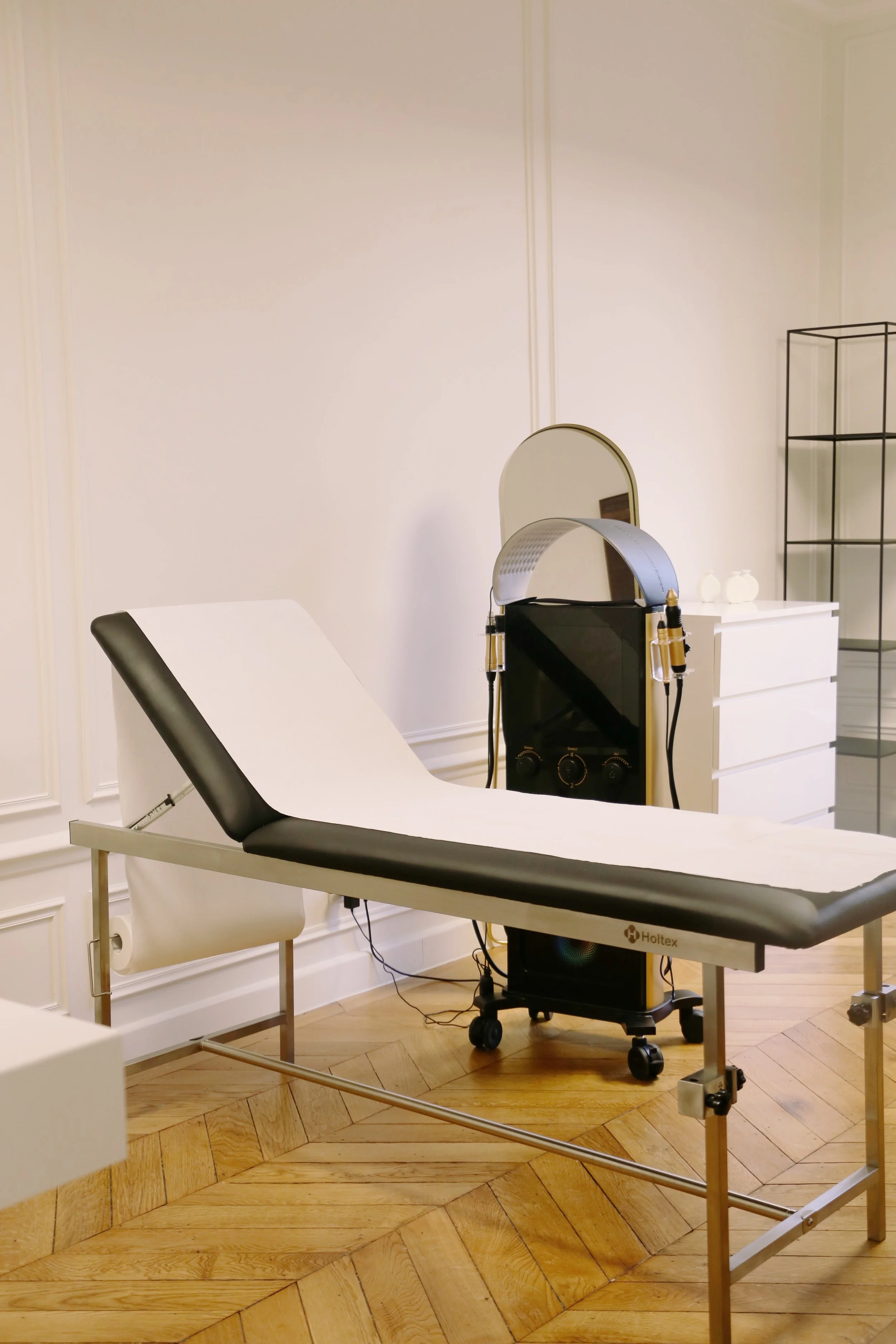 Une salle médicale avec un lit d'examen, un miroir, un appareil médical avec des écouteurs, une armoire blanche et une étagère en métal, dans une pièce à parquet en bois.