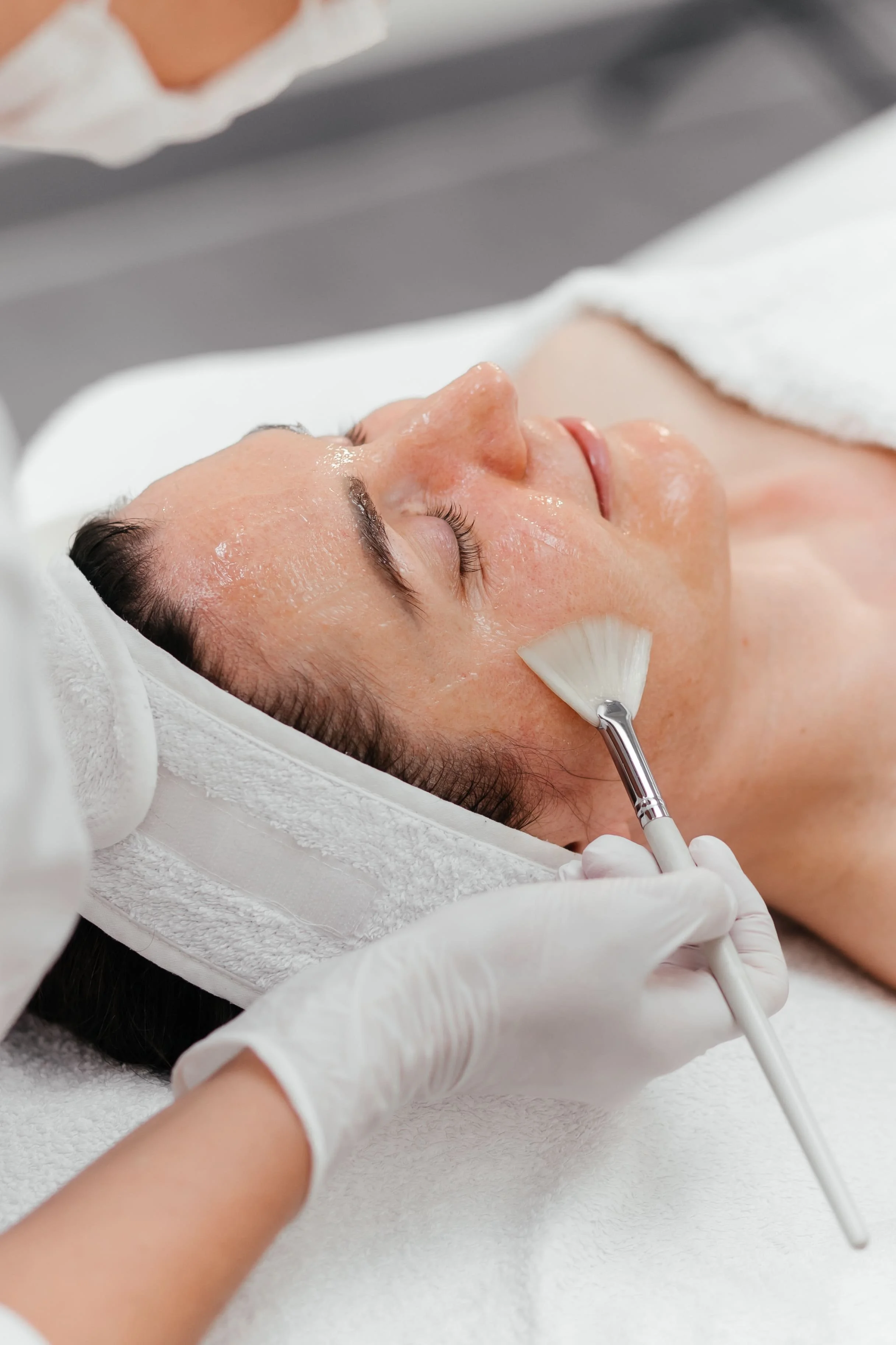 Une femme reçoit un soin du visage dans un spa, allongée sur une table, un professionnel applique un produit à l'aide d'un pinceau.