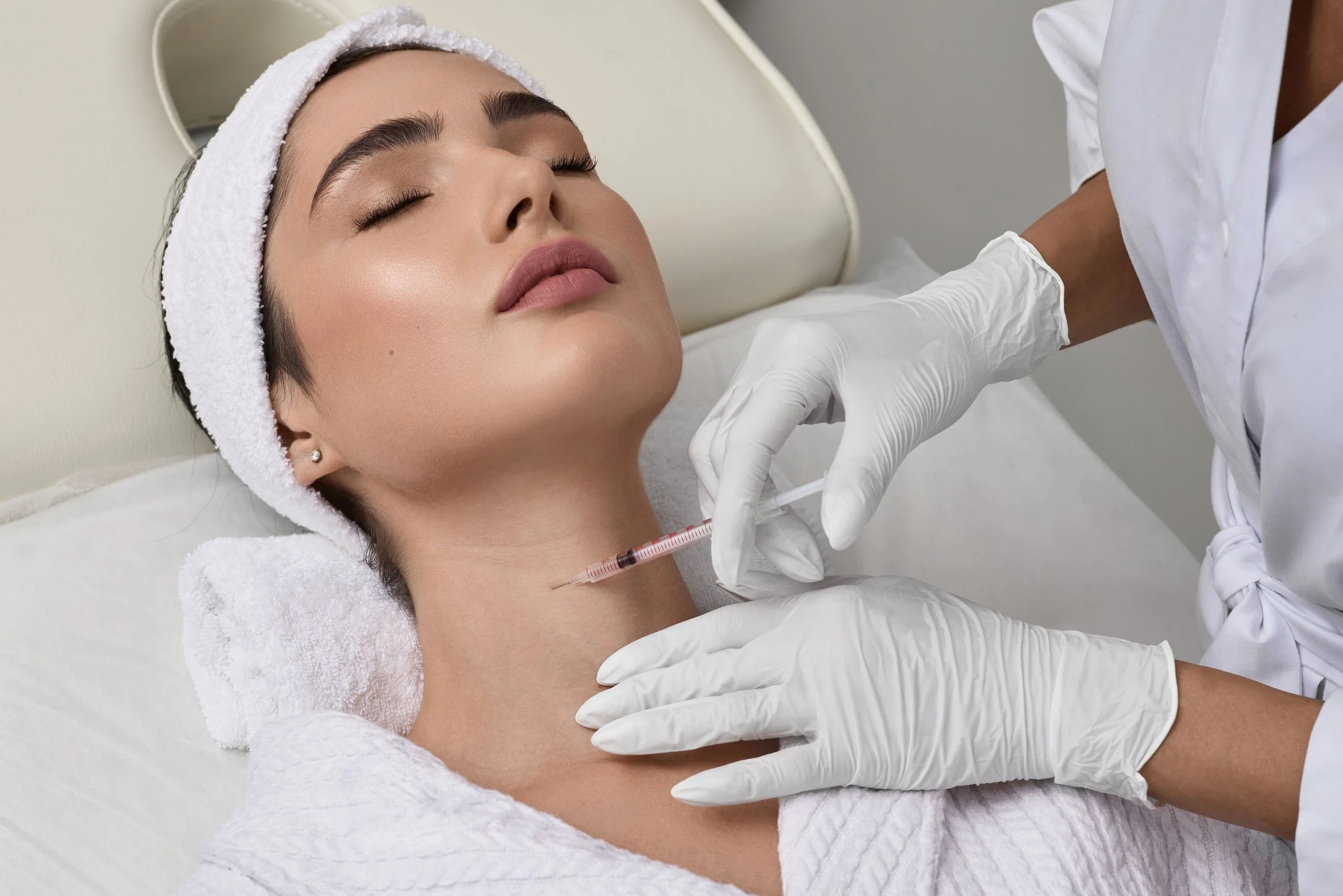 Femme recevant une injection sous le menton dans un contexte médical, patient allongé avec un bandeau blanc, professionnel de santé portant des gants blancs administrant la seringue.