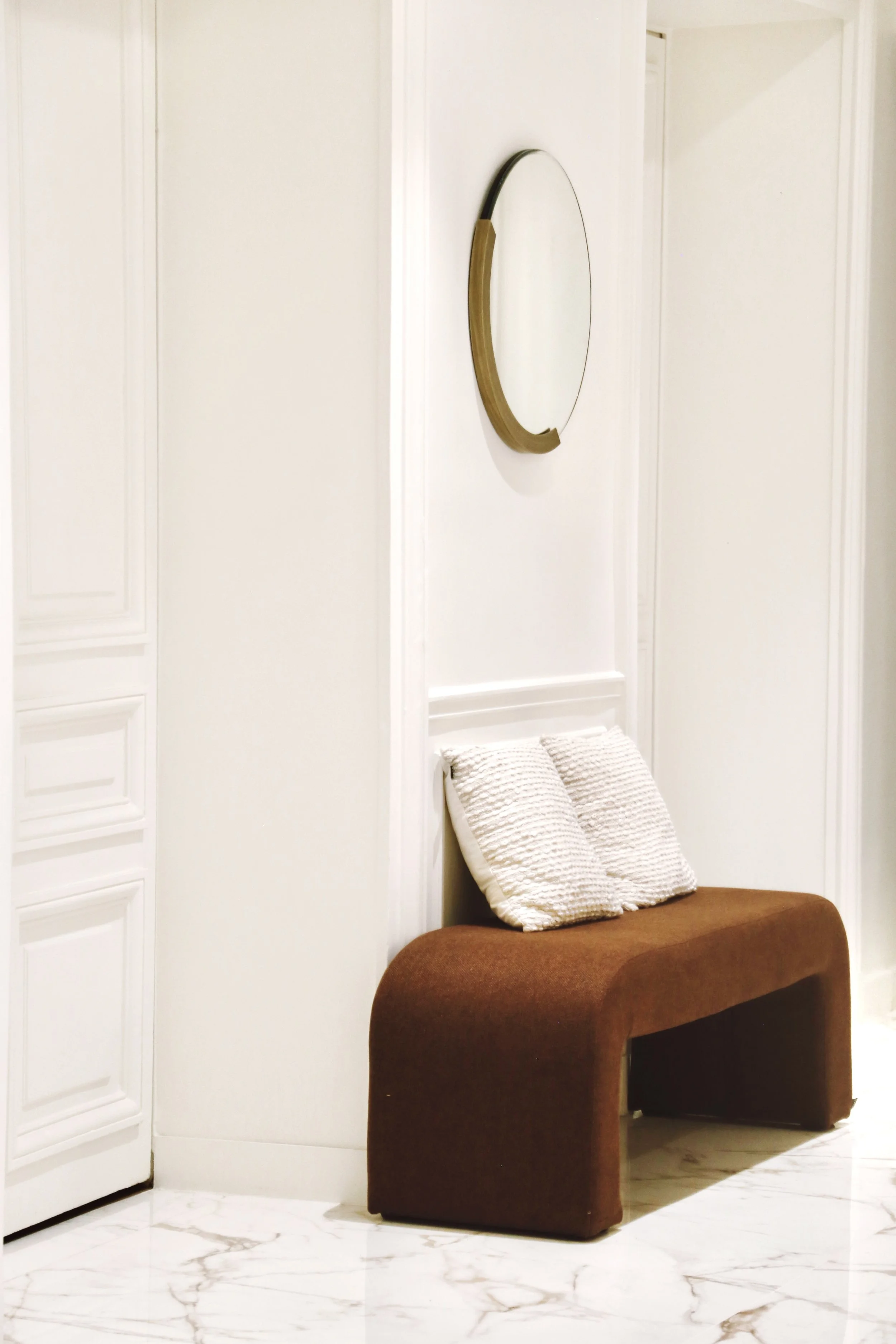 Banquette en tissu marron avec deux coussins beige, miroir rond avec cadre en bois accroché au mur, mur blanc avec moulures, sol en marbre