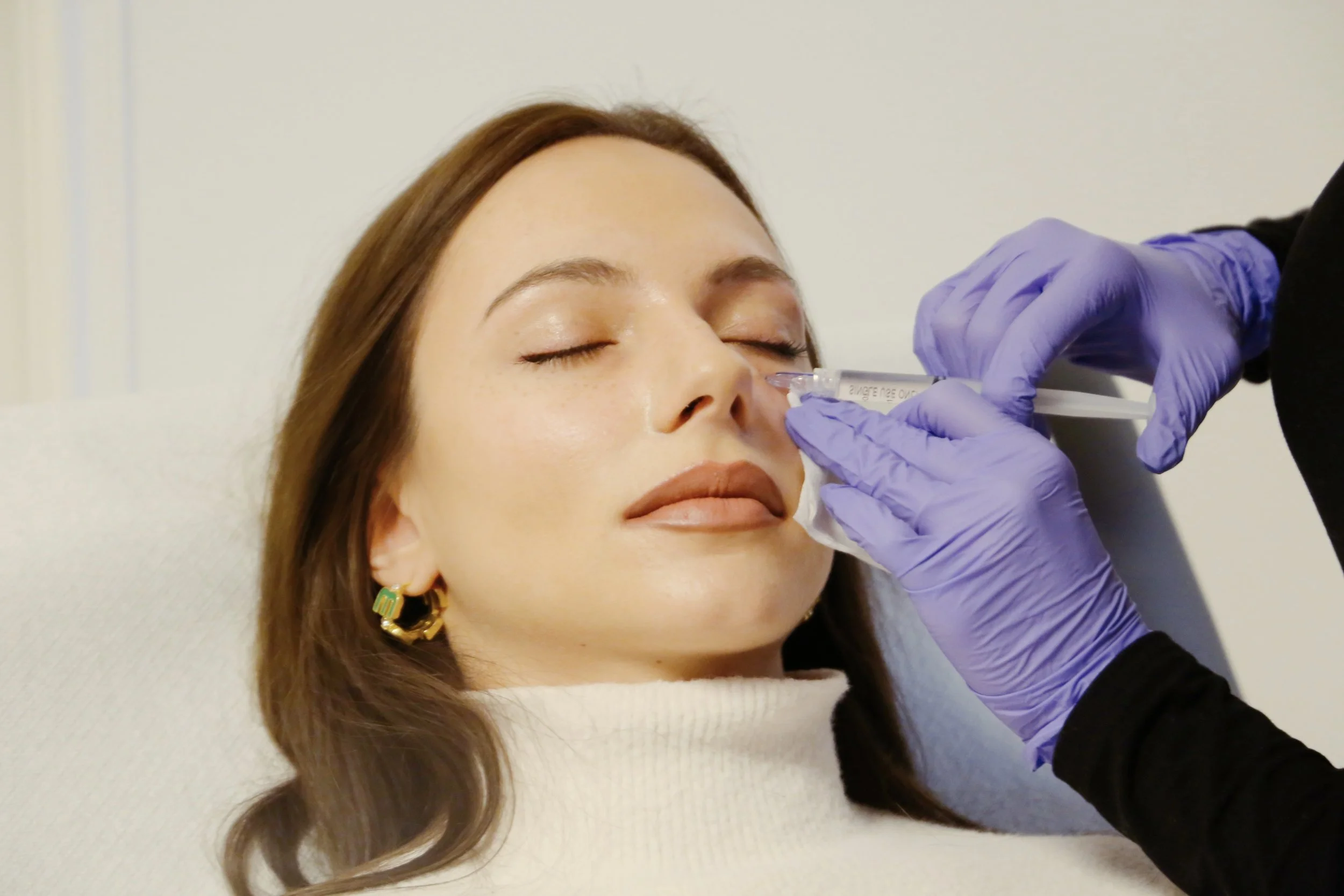 Une femme reçoit une injection injectable dans le nez par un professionnel avec des gants violets, dans un contexte médical ou esthétique.