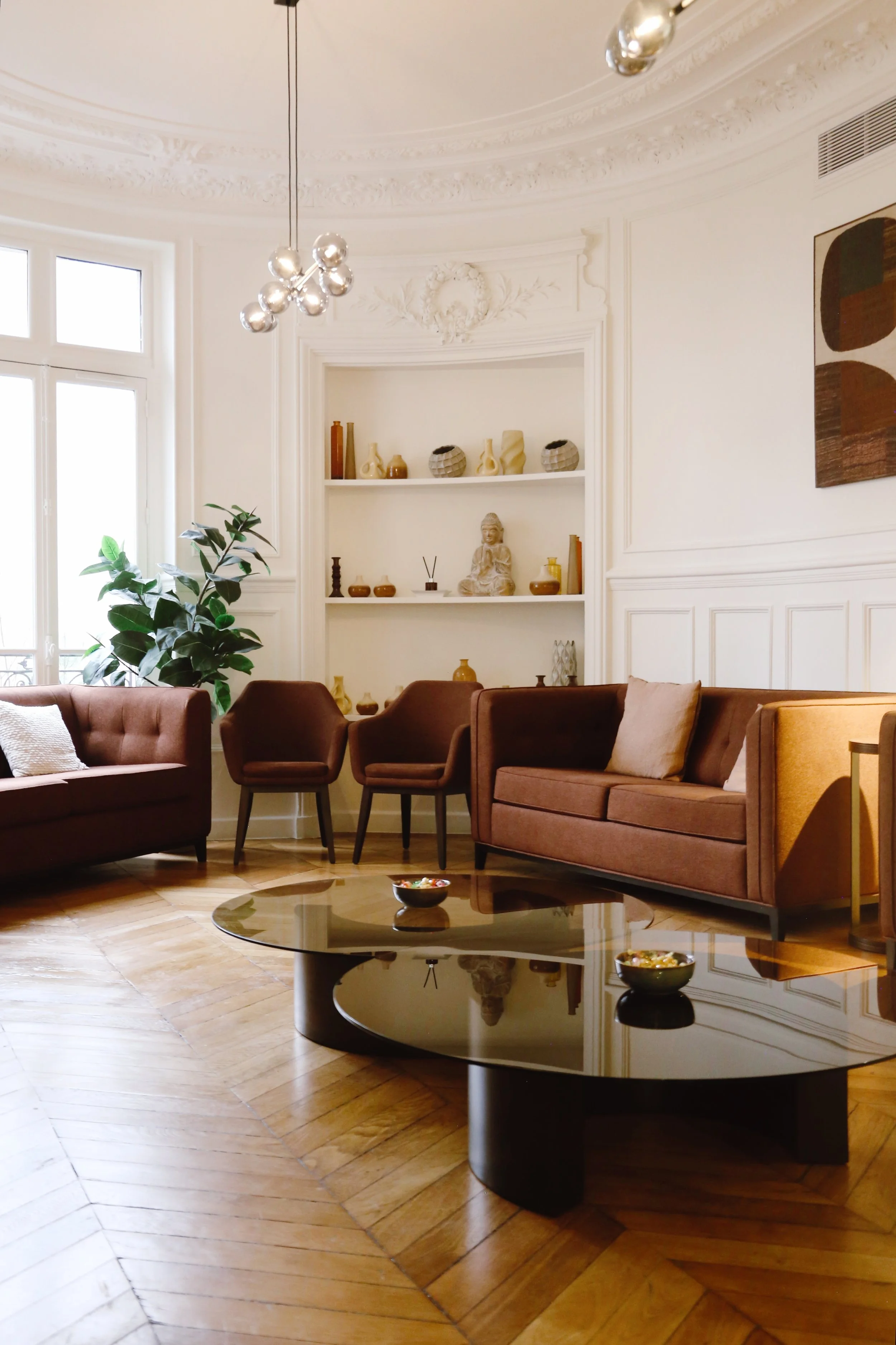 Salon élégant avec canapés marron, chaises et table basse en verre sur parquet en bois, étagère blanche avec décorations, grande fenête avec balcon, plante verte, tableau abstrait, luminaires modernes.