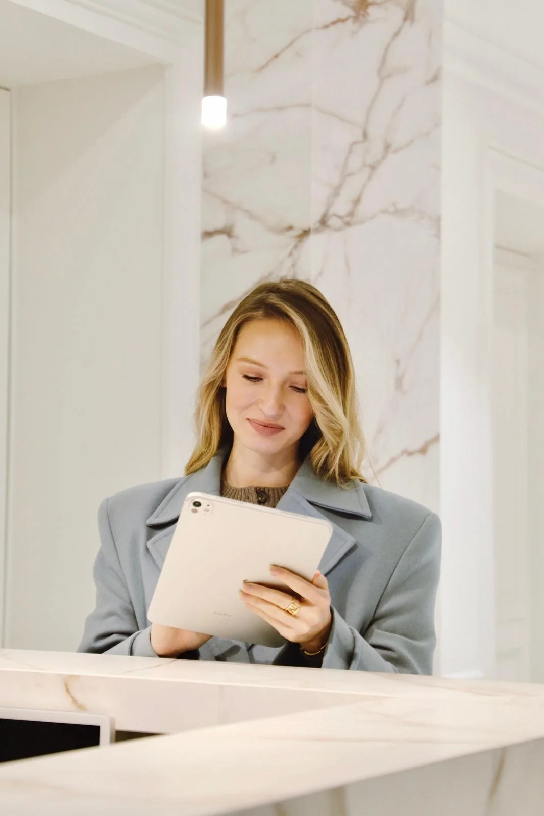 Une femme cheveux blonds portant un manteau gris regarde une tablette blanche dans un intérieur moderne avec des murs en marbre.