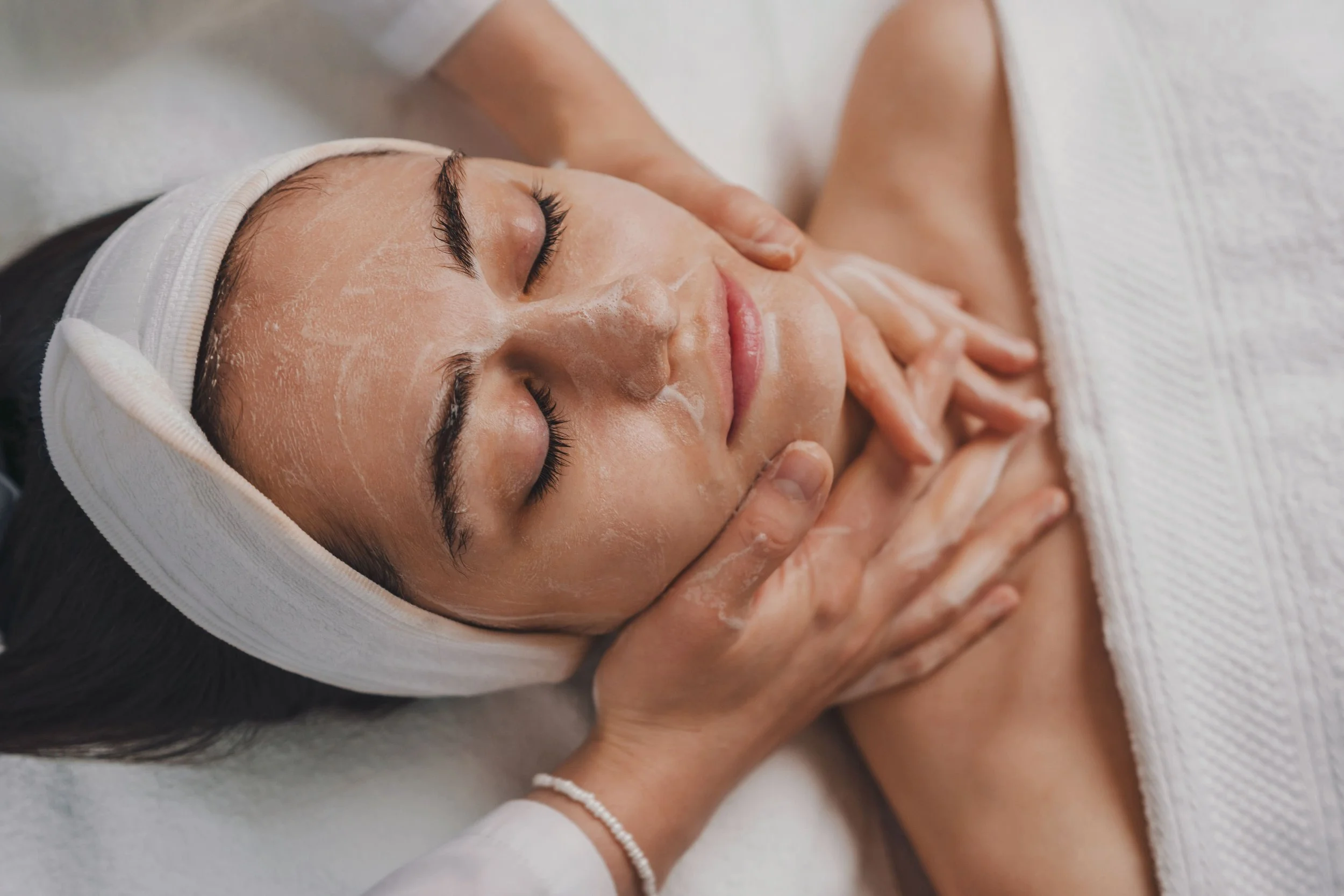 Femme recevant un massage facial dans un spa, avec un masque facial, détendue avec les yeux fermés.