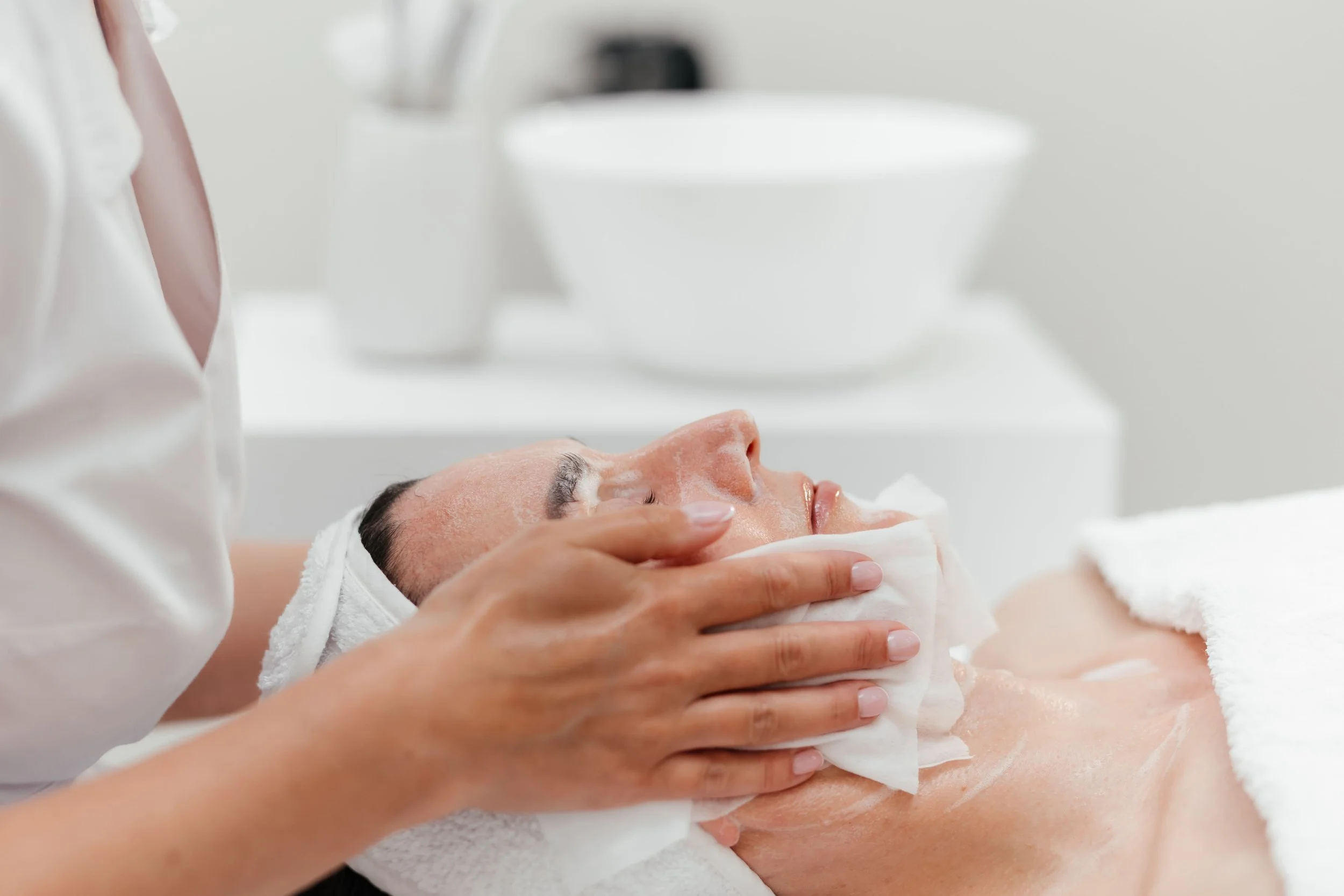 Une personne reçoit un soin du visage dans un spa, avec une autre personne qui essuie son visage avec une serviette. La scène se déroule dans un environnement propre et lumineux.