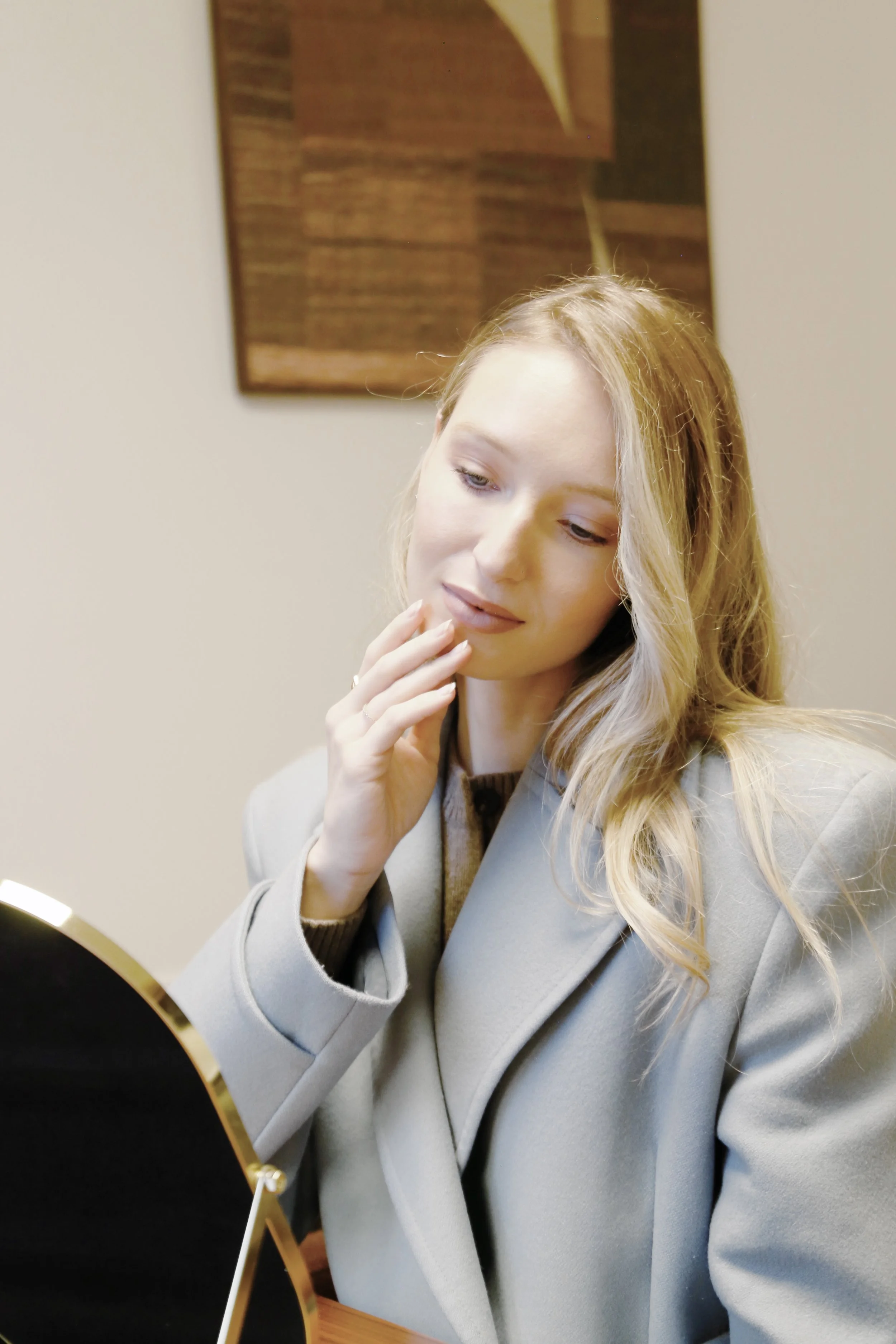 Une femme blonde regarde un miroir avec une expression pensive. Elle porte un manteau gris et est assise à une table.
