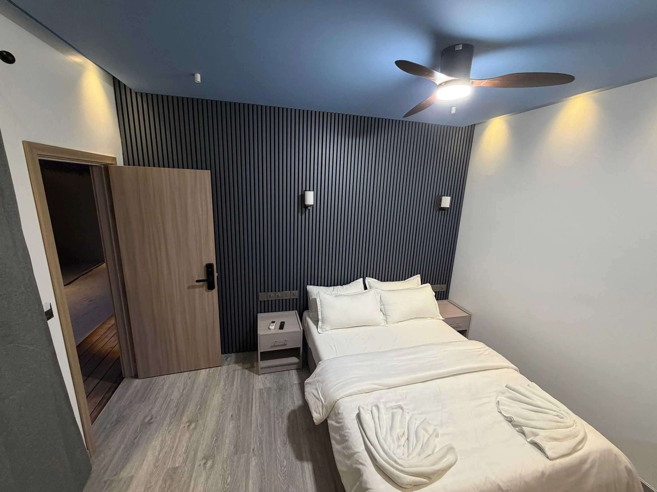 Modern bedroom with a bed, nightstands, and a black slatted accent wall, illuminated by wall-mounted lights and a ceiling fan with wooden blades.