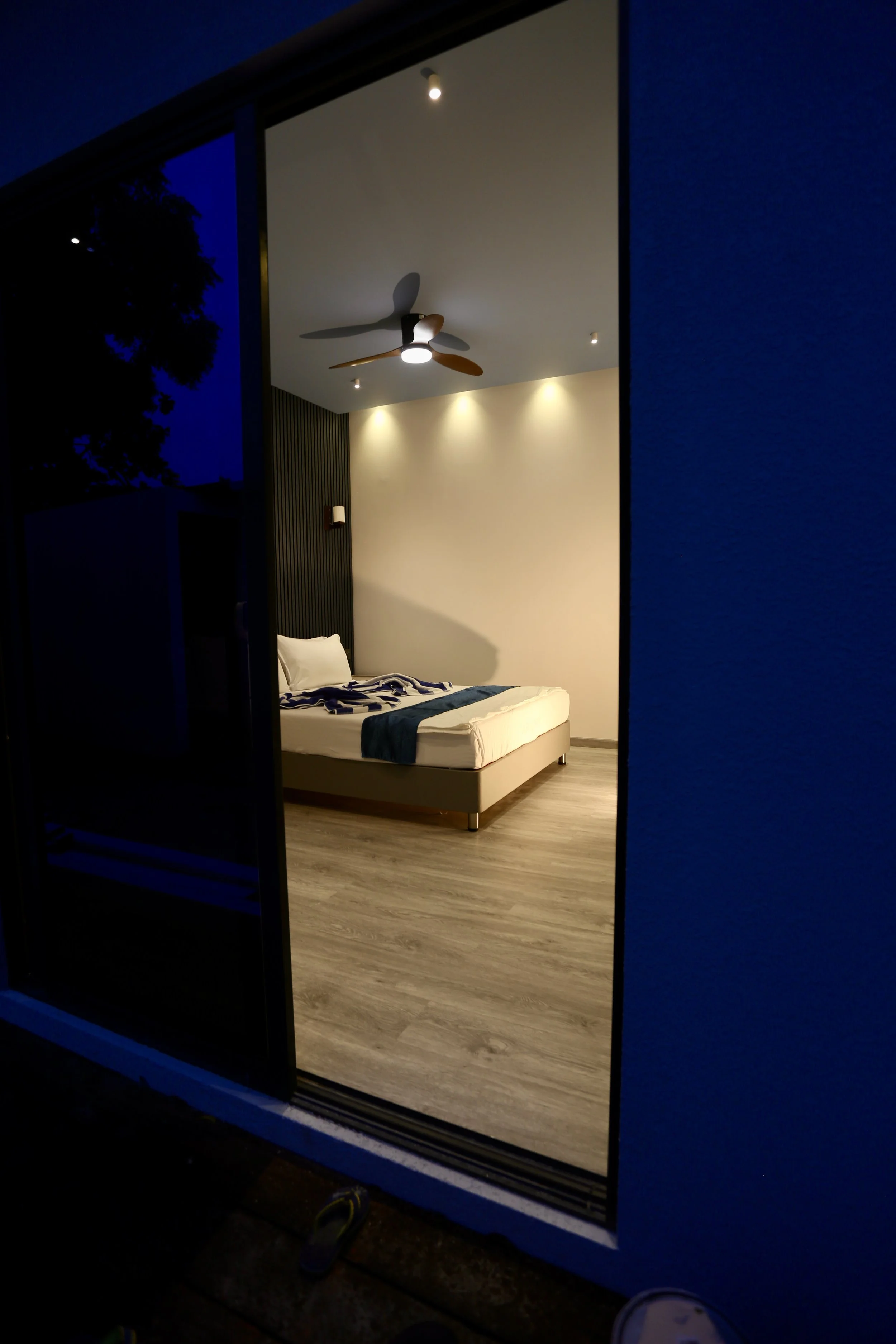 View of a modern, minimalistic bedroom with a bed, ceiling fan, and soft lighting, seen through a sliding glass door at night.