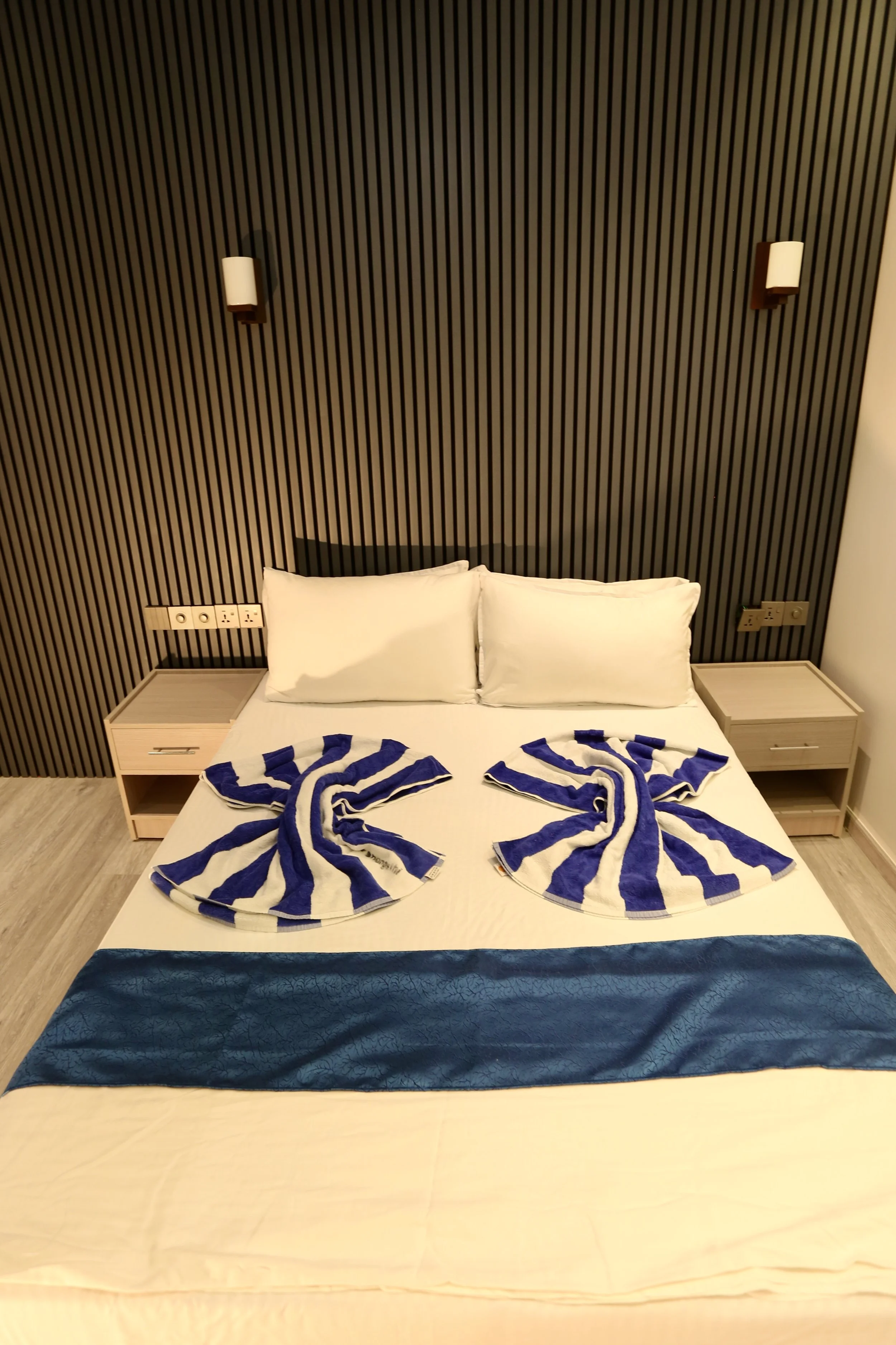A bed with two pillows, decorated with two blue and white towels folded into intricate shapes, in a hotel room with wood-paneled black headboard, bedside tables, and modern lighting.