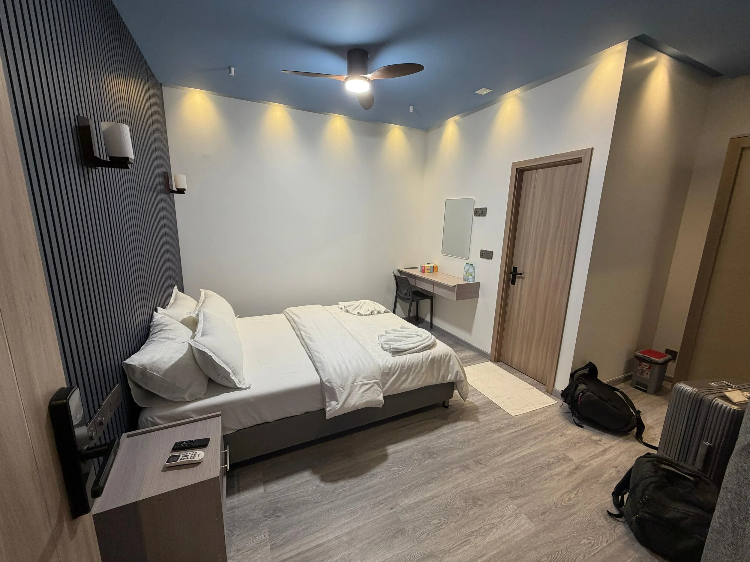 A modern hotel room with a neatly made bed, a nightstand with a remote and phone, a wall-mounted TV, a ceiling fan, a desk with a chair, a mirror, bottles of water, and luggage near the door.