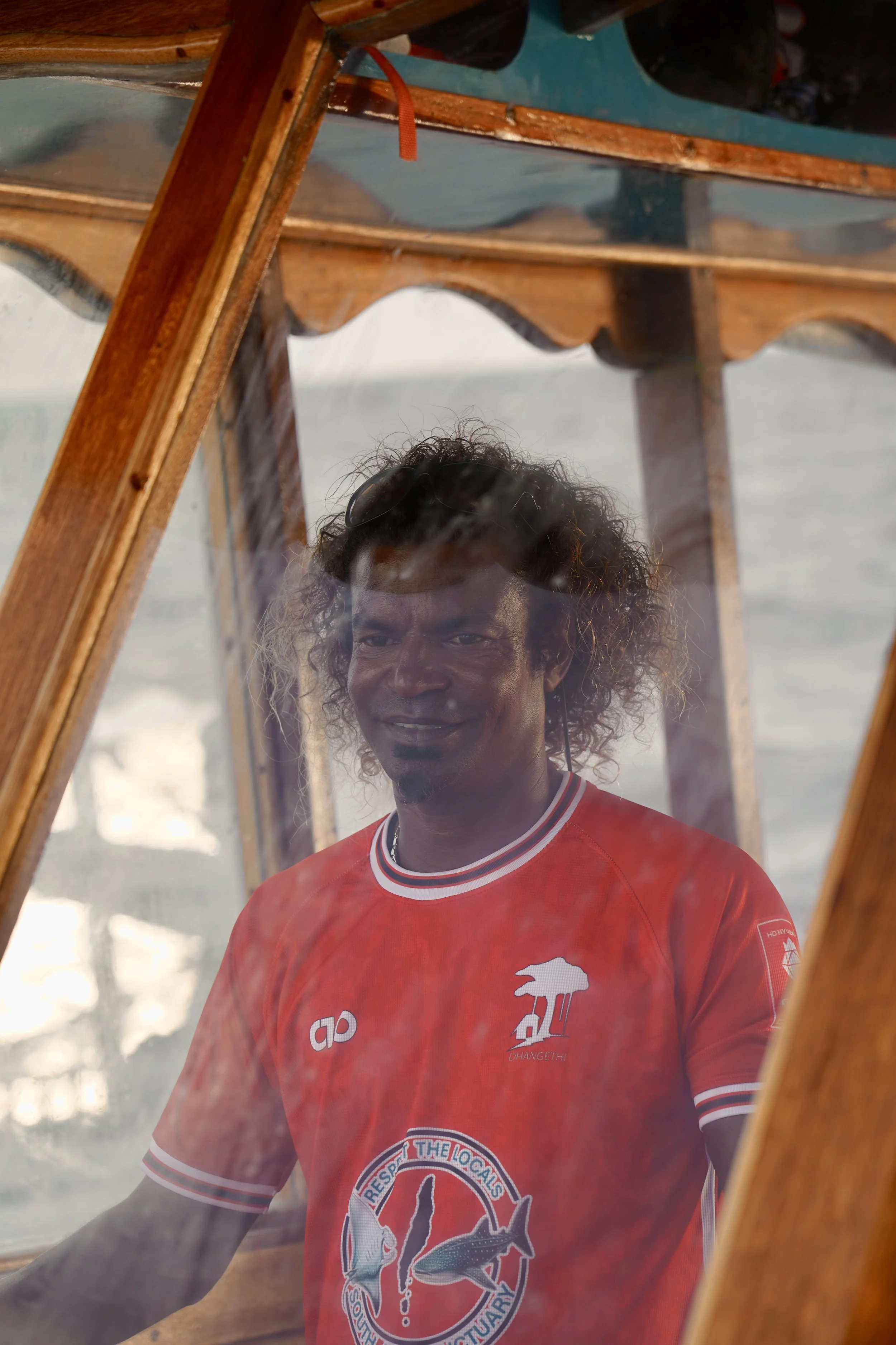 A man with curly hair and sunglasses on his head, wearing a red sports jersey with logos, is seen through a glass window with water reflections, inside a wooden boat.