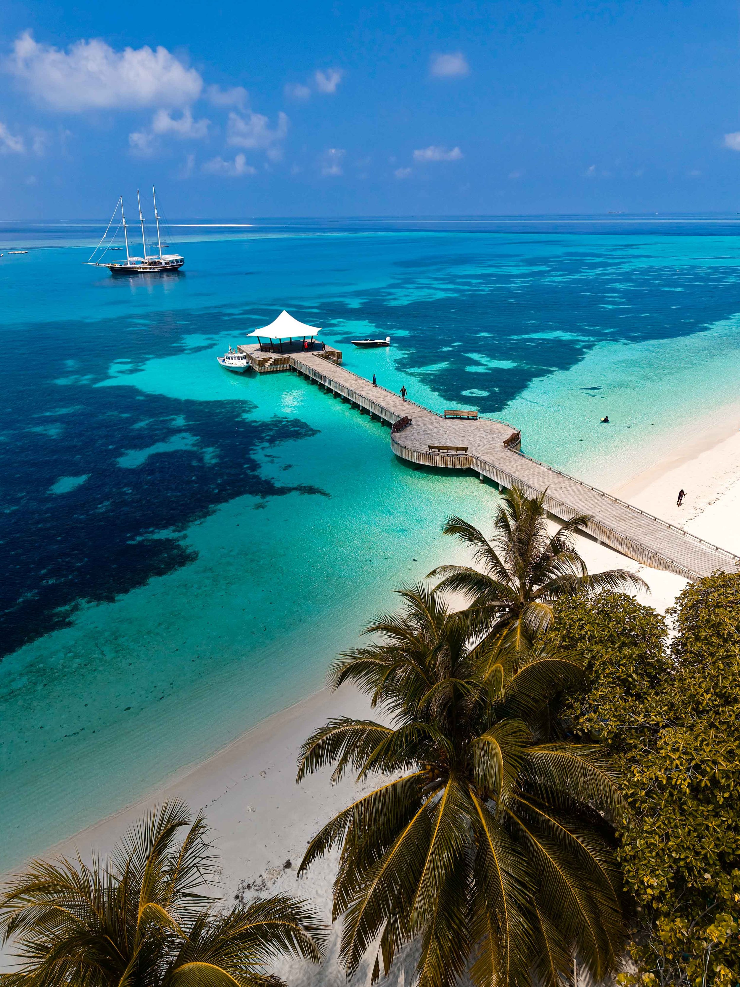 A tropical beach with white sand, palm trees, a pier extending into light blue water, and a large sailing yacht anchored nearby.